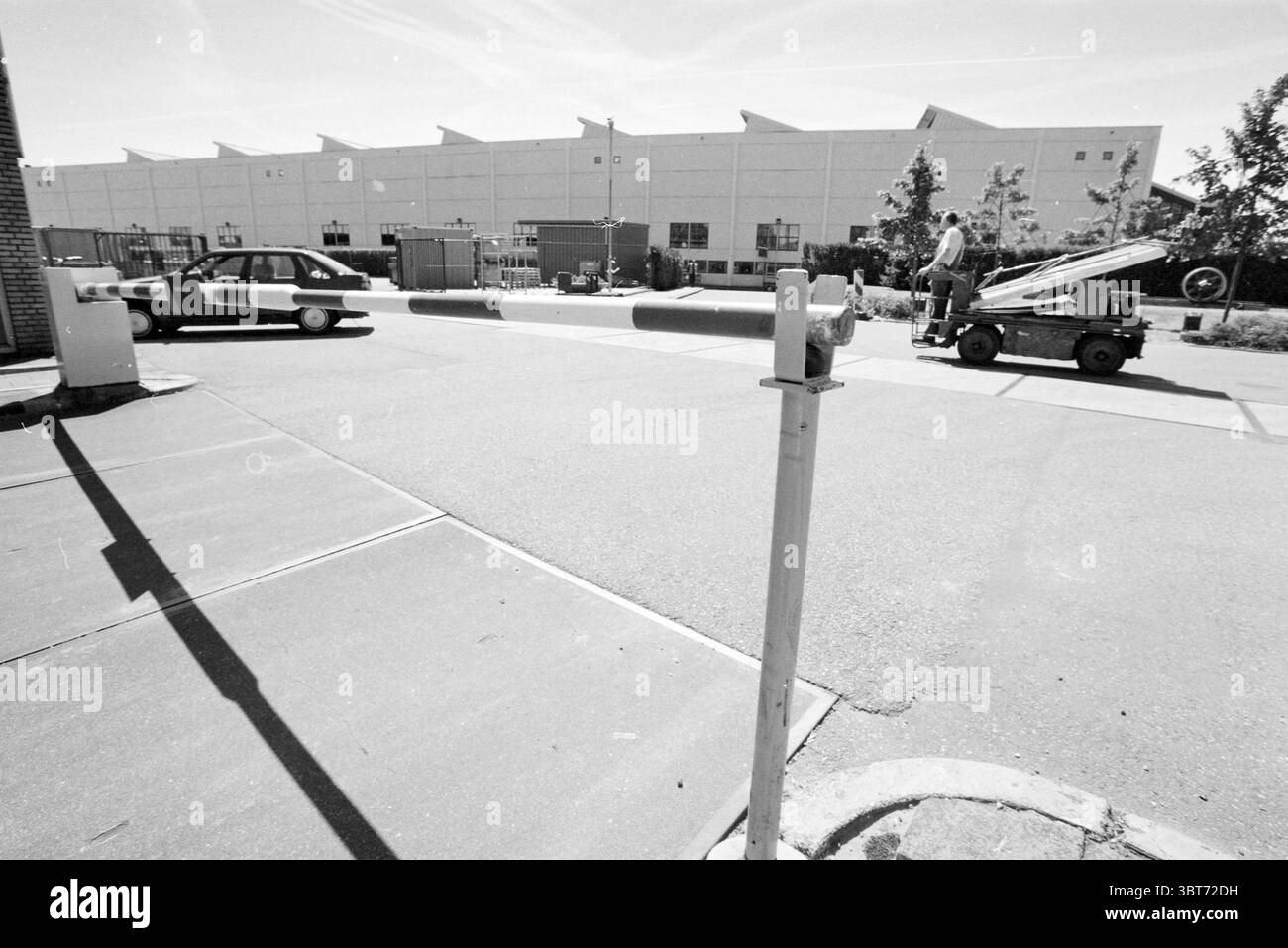 Außenwerkstatt Haarlem the Netherlands, Whizgle News, Dutch Desk, Niederlande, 1950 - 2000 im Jahr 1988. Das Bild enthält diese Themen. In einer monochromatischen Szene wird der Betrachter von einem breiten Bürgersteig begrüßt, der zu einem großen Industriegebäude im Hintergrund führt. Das Äußere des Gebäudes ist in einem gedämpften Ton gestrichen und zeichnet sich durch einen einfachen, boxförmigen architektonischen Stil mit eckigen Dächern aus, die ein starkes Gefühl von Struktur schaffen. Im Vordergrund steht eine Barriere, deren Stange leicht nach links geneigt ist und in einer hellen Farbe gehalten ist, die einen Kontrast zum dunkleren Pflaster bildet. Dieses b Stockfoto