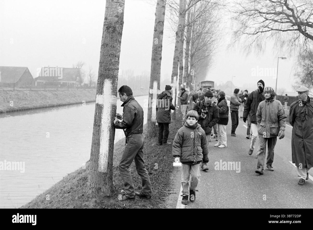 Aktion gegen die 5. Lane Hoofdvaart Haarlemmermeer Hoofdvaart, Whizgle News, Dutch Desk, Niederlande, 1950 - 2000 am 26-11-1993. Das Bild enthält diese Themen. Die Szene zeigt einen nebeligen, bewölkten Tag entlang eines schmalen Kanals. Auf der einen Seite geht eine Gruppe von Menschen auf einem Weg, die sich in einer gemeinsamen Aktivität verstrickt erscheinen. Sie sind in Winterkleidung gebündelt; Jacken in verschiedenen Farben, einige in gedämpften Grau- und Brauntönen, während andere leuchtende Farben wie Blau und Grün tragen. Im Vordergrund läuft ein Junge mit dunklem Mantel und Hut zielgerichtet, hält ein kleines Objekt in der Hand, vielleicht eine Kamera Stockfoto