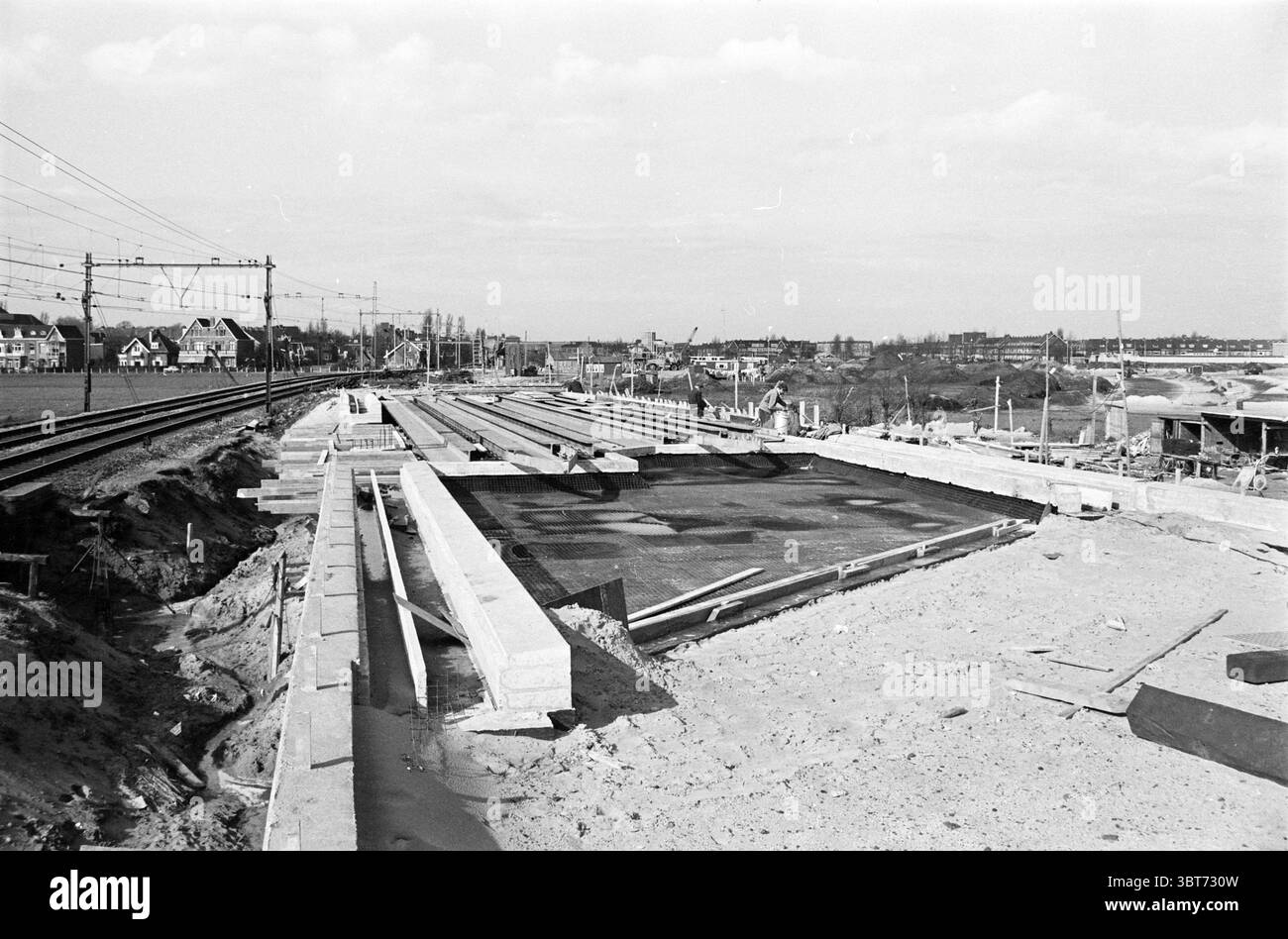 Bau des nördlichen Teils des westlichen Randweges zwischen Bahn und Kleverlaan. Bau von Hochgleis und Viadukten über die neue Straße Haarlem Westelijke Randweg Nederland., Whizgle News, Dutch Desk, Niederlande, 1950 - 2000. Diese Themen werden in der Abbildung angezeigt. Die Szene zeigt eine weitläufige Baustelle, die sich über den Vordergrund erstreckt und eine riesige Fläche mit Roherde und Baumaterialien zeigt. Große, unfertige Betonplatten sind in ordentlichen Reihen angeordnet, was auf den Rahmen einer noch zu vollendenden Struktur hindeutet. Die Textur des Betons wirkt rau, Co Stockfoto