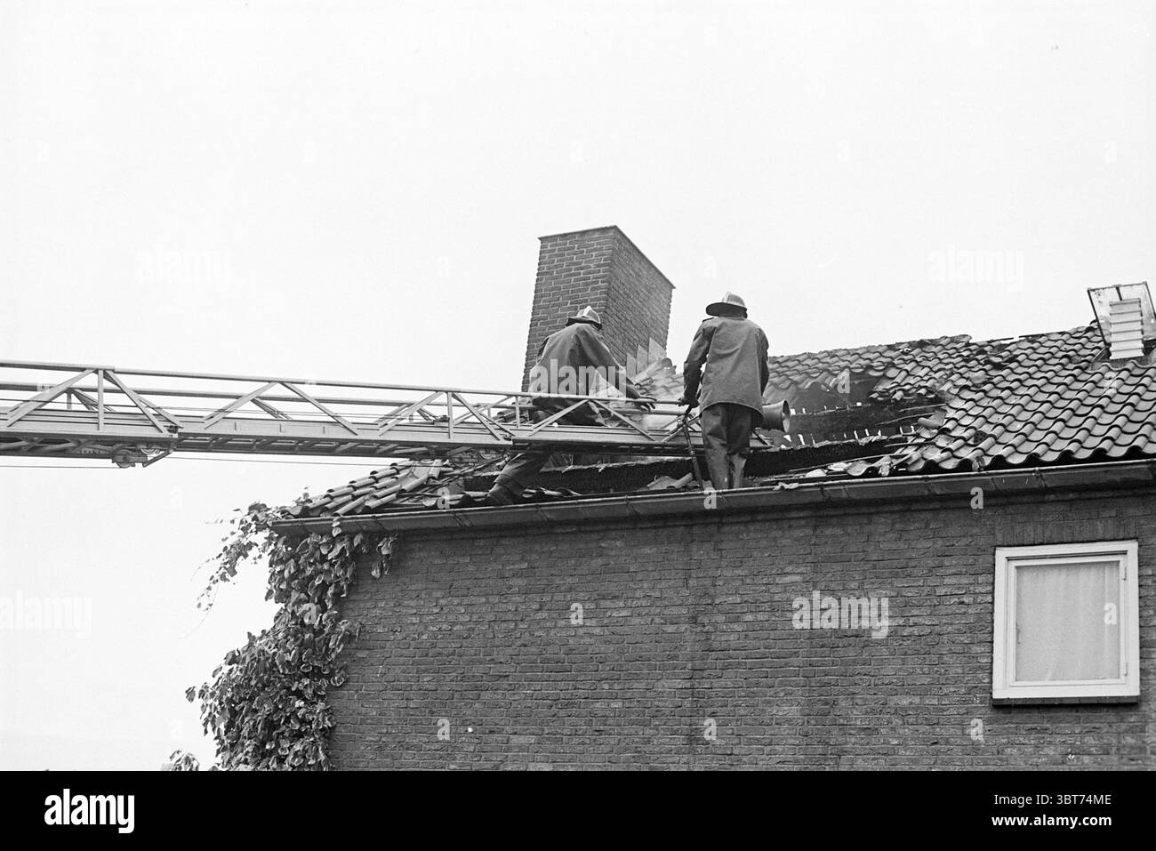 Marke Dr. Schaepmanlaan Heemstede Feuerwehrwehr Heemstede Dr. Schaepmanlaan Niederlande, Whizgle News, Dutch Desk, Niederlande, 1950 - 2000 am 02-08-1974. Diese Themen sind in der Abbildung dargestellt. Auf einem schrägen Dach sind zwei Figuren zu sehen, die an einer dringenden Aufgabe beteiligt sind. Sie tragen dunkle, schwere Kleidung, möglicherweise Schutzausrüstung, was auf den Ernst der Situation hinweist. Eine Person befindet sich in der Nähe des Dachrandes, während die andere etwas hinter dem Dach steht. Beide konzentrieren sich auf einen sichtbar beschädigten Teil des Daches, dessen verkohlte Bereiche auf einen Brand hinweisen. Ein langes Metall Stockfoto