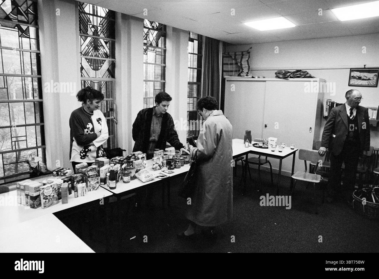 Bazar Good Shepherd Church, Whizgle News, Dutch Desk, Niederlande, 1950 - 2000 am 05-11-1993. Diese Themen werden in der Abbildung angezeigt. In einem geräumigen Zimmer mit großen Fenstern mit Buntglasmotiven treffen sich mehrere Personen um einen langen Tisch. Das natürliche Licht filtert durch das Glas und wirft bunte Muster auf den Boden, obwohl die allgemeine Atmosphäre aufgrund der monochromatischen Töne der Umgebung etwas gedämpft ist. Drei Personen stehen neben dem Tisch, führen Gespräche und untersuchen verschiedene Punkte, die auf der Oberfläche verteilt sind. Der Tisch ist überfüllt mit einem Sortiment o Stockfoto