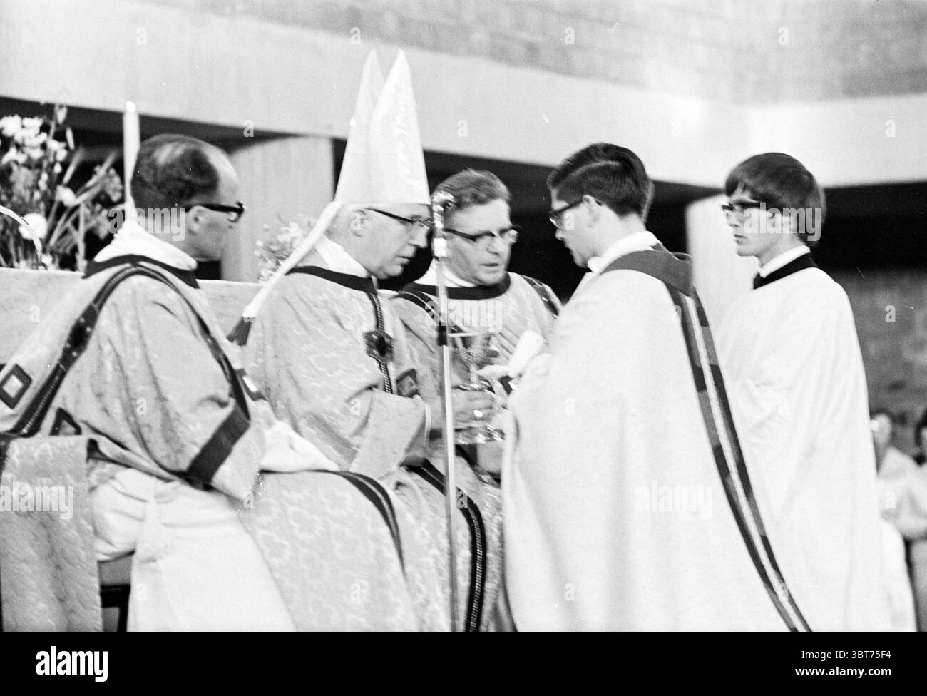 Initiation durch Priester Beverwijk. Initiation, Whizgle News, Dutch Desk, Niederlande, 1950 - 2000 am 05-08-1967. Die Abbildung zeigt diese Themen. Die Szene entfaltet sich in einer feierlichen, religiösen Umgebung, wahrscheinlich einem Ort der Anbetung, geprägt von einem einfachen, aber eleganten architektonischen Design. Das Ambiente ist von Ehrfurcht und Sinn erfüllt. Im Vordergrund steht eine Gruppe von Individuen in traditionellen liturgischen Gewändern. Die amtierenden Figuren tragen aufwendig gestickte Gewänder, wobei eine prominente Person in einer hohen, spitzen Gehrung verziert ist. Die Bademäntel sind hauptsächlich weiß, Stockfoto