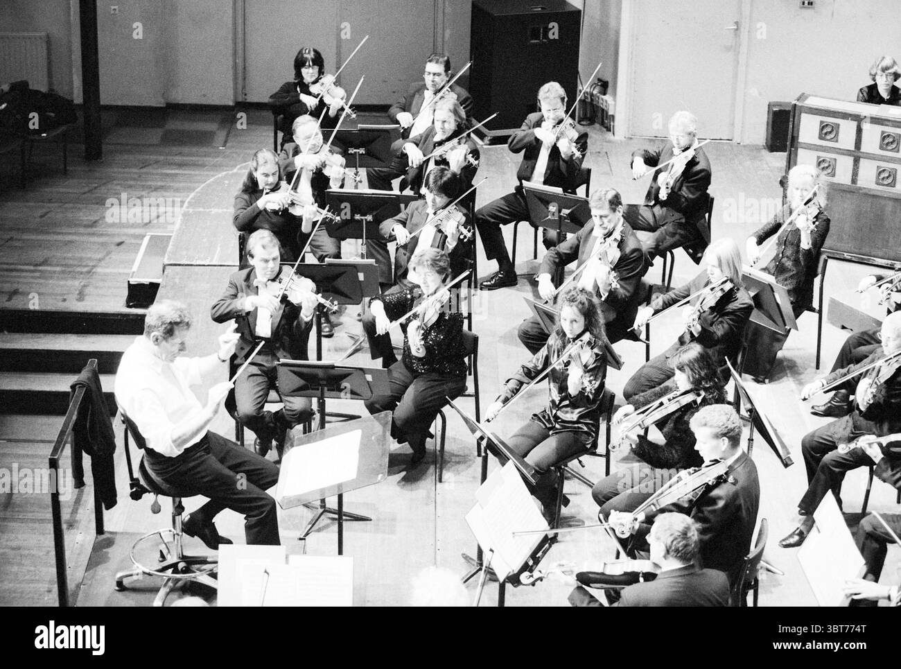 Orchestra NPhO [North Holland Philharmonic Orchestra], Whizgle News, Dutch Desk, Niederlande, 1950 - 2000 am 08-12-1998. Das Bild enthält diese Themen. Die Szene fängt ein Orchester in voller Aufführung in einem geräumigen Konzertsaal mit einem warmen, einladenden Ambiente ein. Die Musiker befinden sich in einer halbrunden Formation um einen zentralen Dirigenten, der sie mit schwungvollen Handbewegungen animiert führt. Das Ensemble besteht aus einer vielfältigen Gruppe, wobei jeder Musiker sich intensiv auf seine Instrumente konzentriert, die von Violinen und Violen bis hin zu Celli und Flöten reichen. Die Musiker tragen formell Stockfoto
