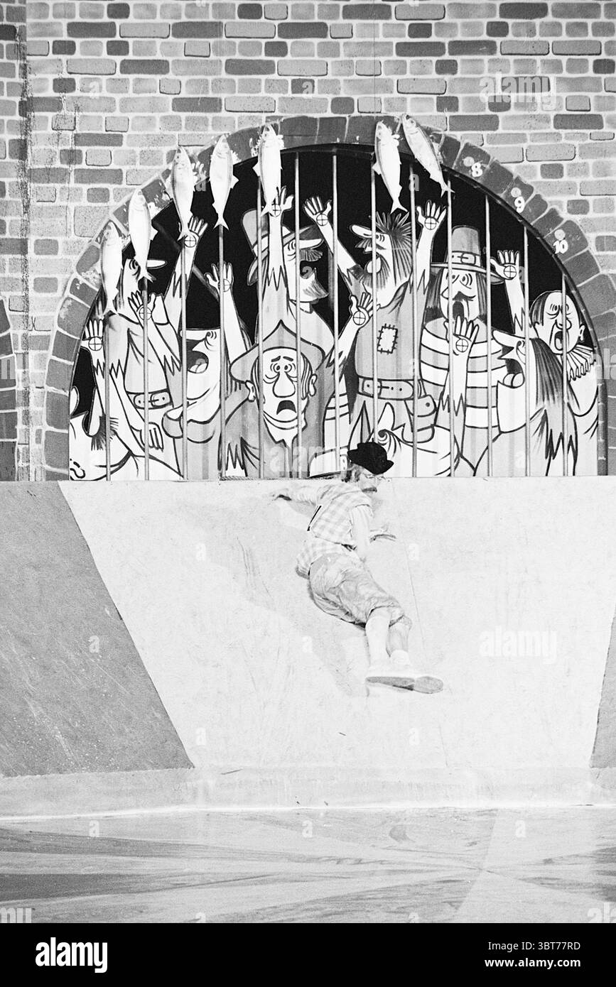 Spiel ohne Grenzen in Leiden Games and Toys, Whizgle News, Dutch Desk, Niederlande, 1950 - 2000 am 18.09.1974. Dies sind die Elemente im Bild. In einer lebhaften Innenumgebung ist ein junger Mensch aktiv, der scheinbar skaten oder einen Stunt aufführt. Die Person, die in hellen Kleidern gekleidet ist, bewegt sich geschickt vor einer Kulisse mit einer großen bogenförmigen Öffnung, die an eine alte Steinmauer mit strukturiertem Backsteinmuster erinnert. Der Bogengang umrahmt ein farbenfrohes Wandbild, das eine skurrile Szene voller überspitzter, cartoonischer Charaktere darstellt. Diese Zahlen sind por Stockfoto