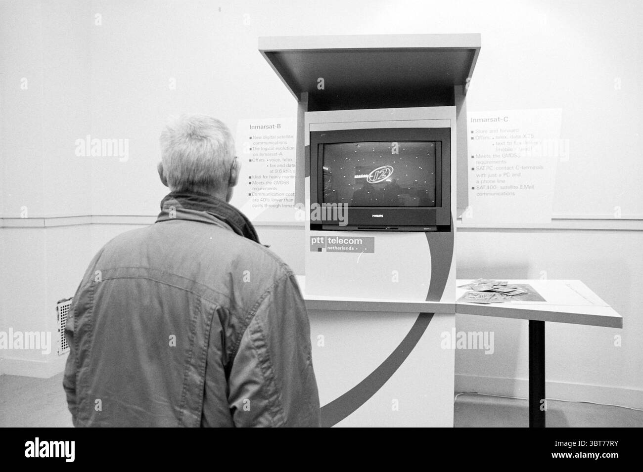 90 Jahre Scheveningen Radio Scheveningen, Whizgle News, Dutch Desk, Niederlande, 1950 - 2000 am 28-10-1994. Das Bild enthält diese Themen. Die Szene zeigt eine Person, die vor einem Kiosk steht und ein schlankes, modernes Design hat. Die Person steht vor einem Monitor, der eine lebendige Grafik mit Himmelsbildern zeigt, einschließlich Sternen und möglicherweise einem Raumschiff. Die Person trägt eine Jacke in neutraler Farbe, die eine dezente Palette andeutet, die sich mit den hellen Bildern auf dem Bildschirm abhebt. Der Kiosk selbst ist überwiegend weiß mit roten Akzenten, was eine moderne Gebühr bietet Stockfoto