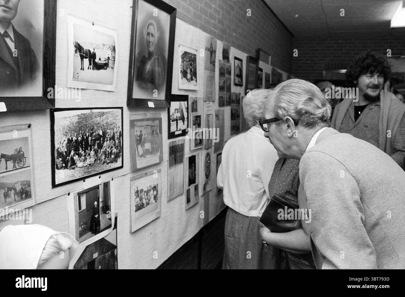 Eröffnung Ausstellung 100 Jahre Seebad Wijk aan Zee Eröffnungen Inbetriebnahme Ausstellungen Wijk aan Zee, Whizgle News, Dutch Desk, Niederlande, 1950 - 2000 am 07-07-1980. Dies sind die Elemente im Bild. Die Szene spielt in einem warm beleuchteten Innenraum, der einem Gemeindesaal oder einer Galerie ähnelt. Die Wände sind mit einer Collage aus verschiedenen gerahmten Fotografien verziert, die Schwarz-weiß-Bilder zeigen, die ein Gefühl von Nostalgie hervorrufen. Die Fotografien zeigen eine Reihe von Themen, von historischen Ereignissen bis hin zu offenen Porträts, die der Atmosphäre Tiefe verleihen. Im Vordergrund steht eine Gruppe von intellektuellen Menschen Stockfoto
