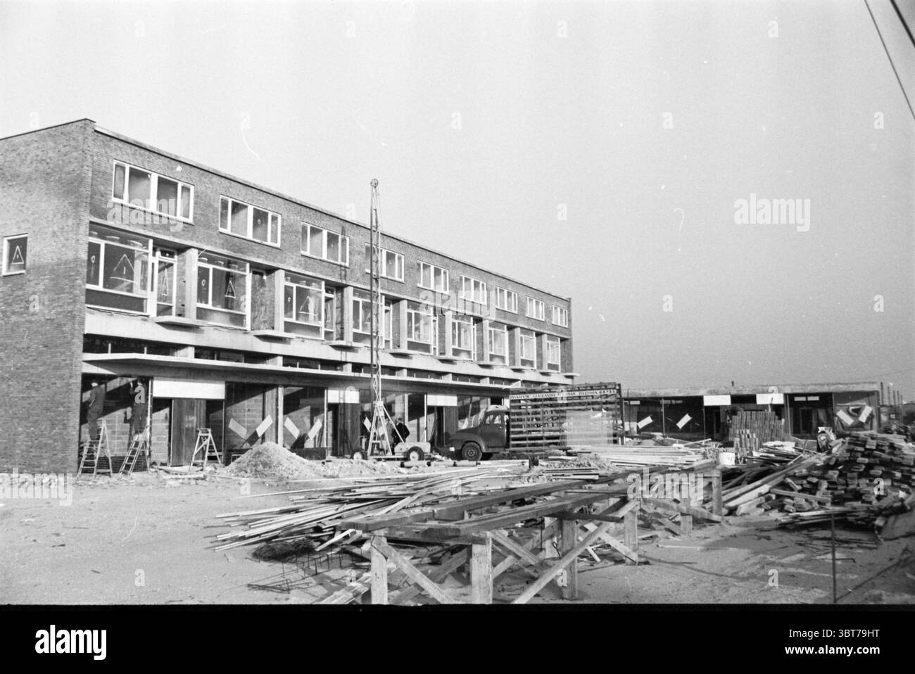 Neuer Komplex Schalkwijk Häuser und Gebäude, Whizgle News, Dutch Desk, Niederlande, 1950 - 2000 am 26-01-1961. Diese Themen sind in der Abbildung dargestellt. Die Szene zeigt eine Baustelle mit Schwerpunkt auf einer Reihe neu errichteter Gebäude. Die Architektur verfügt über mehrere Ebenen, von denen jede mit Reihen von Fenstern verziert ist, die nicht verziert sind und funktionell wirken, was ein zeitgenössisches Design andeutet. Das Äußere des Gebäudes zeichnet sich vor allem durch einen gedämpften Ziegelton aus, der durch die leicht reflektierenden Glasscheiben ergänzt wird. Im Vordergrund sind verschiedene Baumaterialien verstreut Stockfoto