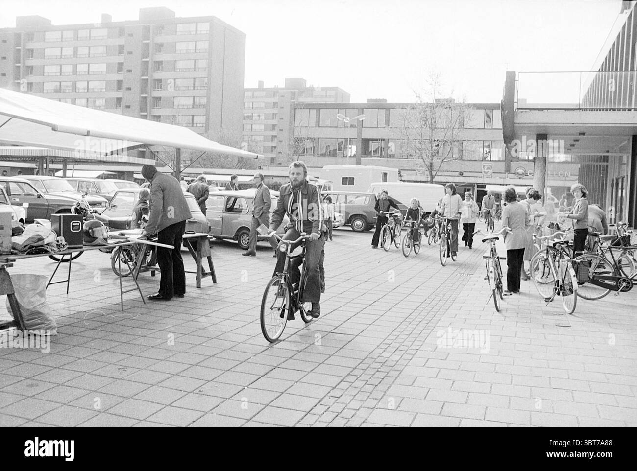 Beginnen Sie den Radtag Beverwijk und IJmuiden Radwege Radfahrer Start, Whizgle News, Dutch Desk, Niederlande, 1950 - 2000 am 13-05-1978. Diese Themen sind in der Abbildung dargestellt. Die Szene fängt ein lebhaftes urbanes Ambiente mit einem plaza voller Aktivität ein. Mehrere Personen sind in verschiedenen Aufgaben tätig und schaffen ein Gefühl von Gemeinschaft und Bewegung. Im Vordergrund steht ein Mann auf einem Fahrrad, dessen Ausdruck fokussiert ist, während er durch die Menge navigiert. Um ihn herum laufen mehrere Personen entweder zu Fuß oder stehen an Fahrrädern, einige scheinen sich zu unterhalten oder Gegenstände auf Tischen zu untersuchen. Die Atmosphäre Stockfoto