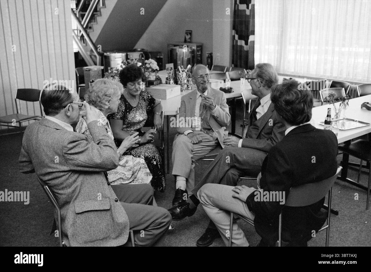 25 Jahre Jubiläum von Herrn H. Beyk Jubiläumsjubiläen VNU Association of Dutch Publishers, Whizgle News, Dutch Desk, Niederlande, 1950 - 2000 am 13.06.1980. Dies sind die Elemente im Bild. Die Szene zeigt eine Zusammenkunft von sechs Personen, die in einem Halbkreis sitzen und sich in Gesprächen befinden. Sie wirken entspannt und animiert, was auf eine freundliche Atmosphäre hindeutet. Das Ambiente ist ein geräumiger Innenbereich mit weichem Teppich und minimalistischer Einrichtung. Im Hintergrund lassen große Fenster natürliches Licht ein, das einen warmen Glanz erzeugt und den Bereich beleuchtet. Helle Vorhänge kreatisieren Stockfoto