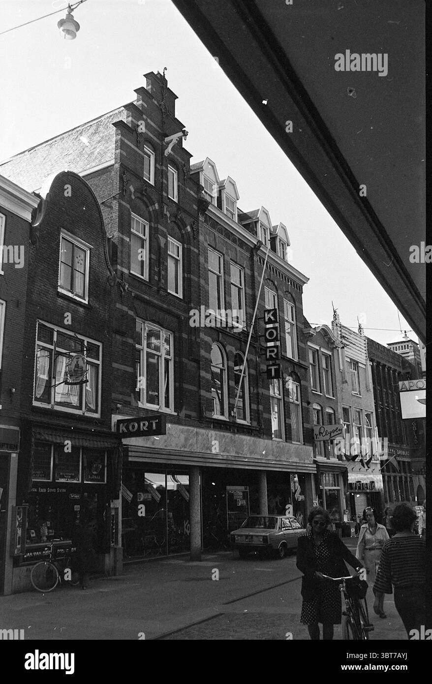 Außenansicht Fa. Kort Grote Houtstraat Haarlem Außengeschäfte und Kioske Haarlem Grote Houtstraat Niederlande, Whizgle News, Dutch Desk, Niederlande, 1950 - 2000 am 16-06-1976. Dies sind die Themen im Bild. Die Szene fängt ein lebhaftes urbanes Ambiente mit einem prominenten Gebäude mit komplexen architektonischen Details ein. Die Struktur weist sowohl spitze als auch geschwungene Elemente entlang einer dunklen Fassade auf, die eine Mischung von Stilen offenbart, die auf historische Einflüsse hinweisen. Große Fenster erstrecken sich über die oberen Stockwerke und erlauben einen Blick in den Innenraum, während das Erdgeschoss durch eine Wide gekennzeichnet ist Stockfoto