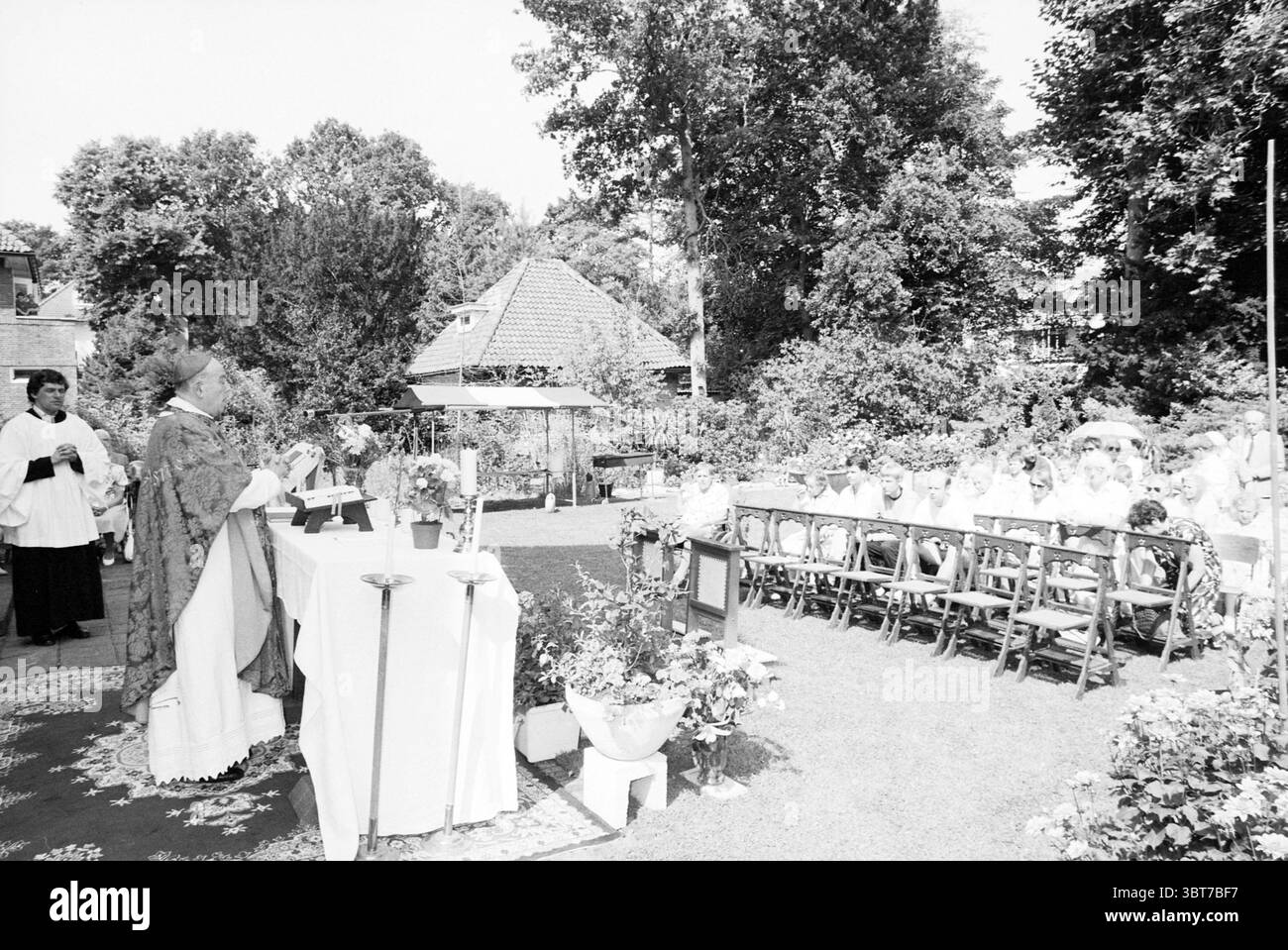 Katholischer Gottesdienst im Freien im Garten der Villa, Whizgle News, Dutch Desk, Niederlande, 1950 - 2000 am 12-08-1990. Dies sind die Themen im Bild. Die Szene spielt im Freien, unter einer hellen Sonne, die einen üppig grünen Garten beleuchtet. Es findet eine Zusammenkunft statt, die um einen großen Tisch mit weißem Leinen gedeckt ist. An der vordersten Front führt ein in traditionellen Gewändern gekleidetes Geistliches eine Zeremonie durch, wobei seine Hände in einer Geste des Segens oder Gebets erhoben werden. Er steht hinter dem Tisch, der mit verschiedenen Blumenarrangements verziert ist und einen lebhaften Kontrast zum Weißen bildet Stockfoto