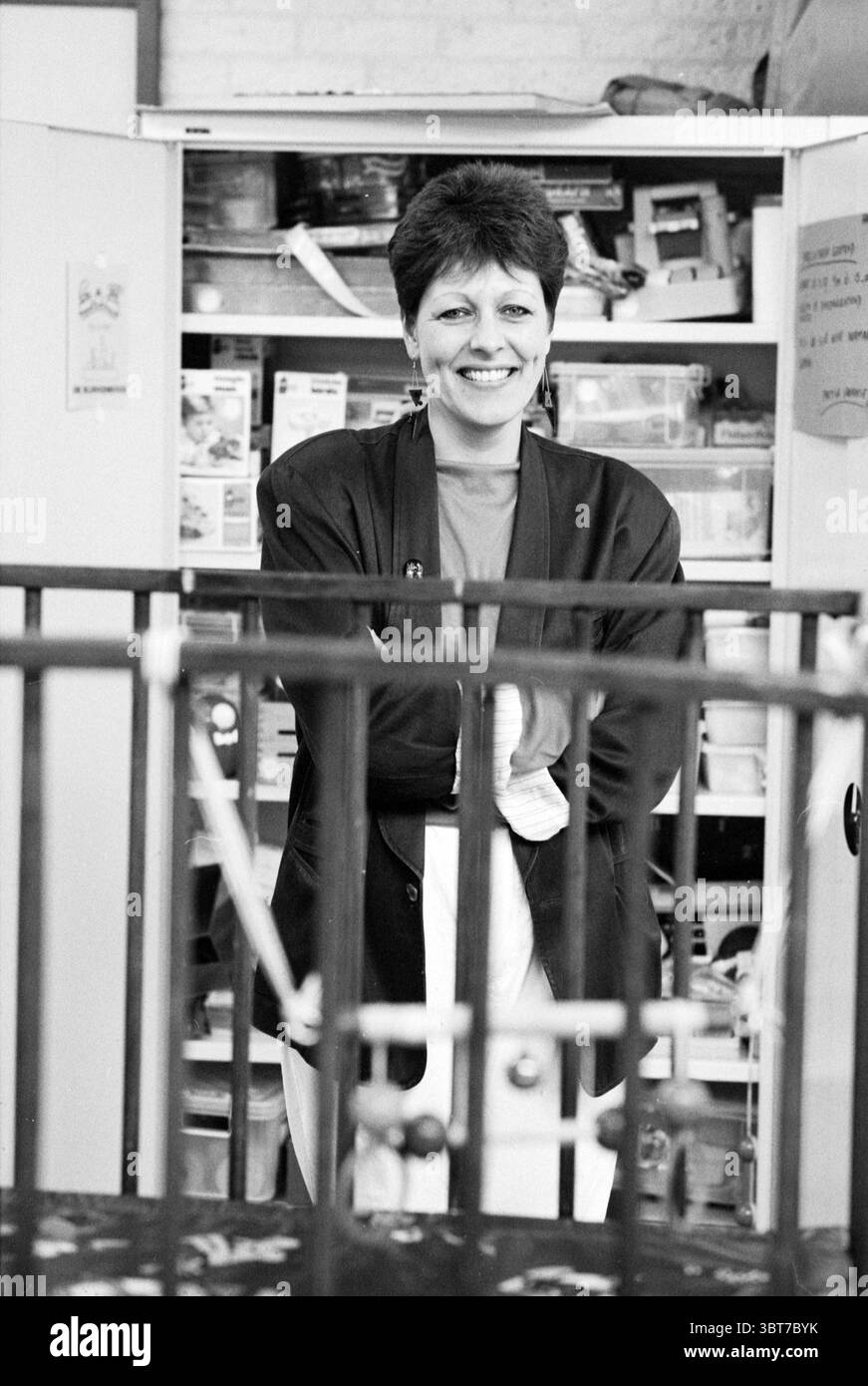 Hoofddorp Spielzeugbibliothek Hoofddorp the Netherlands, Whizgle News, Dutch Desk, Niederlande, 1950 - 2000 am 19-06-1991. Das Bild enthält diese Themen. In der Szene steht eine Frau lächelnd in einem gut beleuchteten Raum, der eine warme und einladende Atmosphäre vermittelt. Sie hat kurze, dunkle Haare und trägt einen schwarzen Blazer über einem hellroten Oberteil, das einen auffälligen Kontrast zu den gedämpften Farben des Hintergrunds bietet. Hinter ihr befinden sich teilweise sichtbare Regale mit verschiedenen Objekten, die auf einen Arbeitsplatz oder einen Bastelbereich hinweisen, der wahrscheinlich mit Werkzeugen und Materialien überfüllt ist. Die Einstellung ist noch organisiert Stockfoto