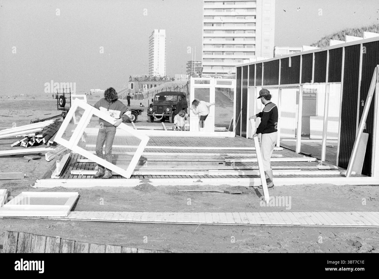 Bau Strandzelte Zandvoort Strand und Strände Zandvoort, Whizgle News, Dutch Desk, Niederlande, 1950 - 2000 am 08-02-1989. Diese Themen werden in der Abbildung angezeigt. In einer Küstenlandschaft wird eine Gruppe von Einzelpersonen auf einer Plattform aus Holzdielen mit Bauarbeiten beschäftigt. Die Farbpalette wird von gedämpften Grau- und Weißtönen dominiert, die ein monochromatisches Schema widerspiegeln, das für die Szene typisch ist. Im Vordergrund stehen drei Personen, die aktiv arbeiten: Zwei Männer und eine Frau. Der erste Mann steht und hält einen großen, rechteckigen Holzrahmen, der sich darauf konzentriert, ihn mit der Struktur auszurichten. Stockfoto
