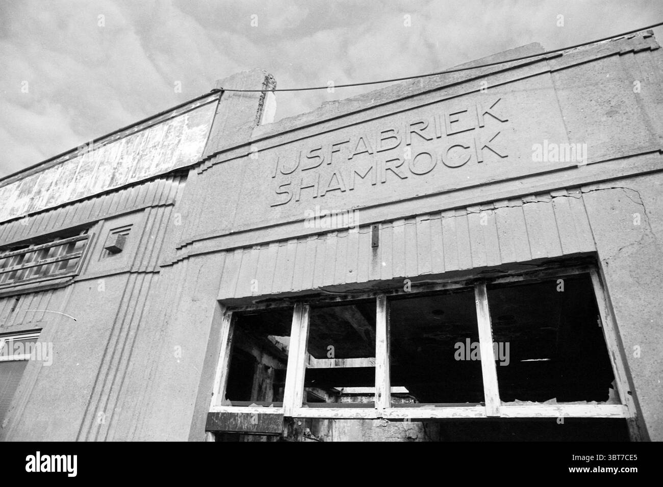 Diverse Hafengegend, Whizgle News, Dutch Desk, Niederlande, 1950 - 2000 am 29-03-1993. Dies sind die Elemente im Bild. Die Szene präsentiert ein industrielles Umfeld, das ein Gefühl von Verfall und Verlassenheit vermittelt. Die Struktur besteht aus rauem Beton und zeigt deutlich einen Namen, der in die Oberfläche eingeätzt ist, was auf ein einst aktives Geschäft hindeutet. Das Gebäude verfügt über einen ausgeprägten architektonischen Stil mit scharfen Linien und einer strukturierten Fassade, die typisch für ältere Industriedesigns ist. Große Fenster sind vorhanden, aber viele von ihnen sind kaputt oder fehlen, was die Leere des Innenraums offenbart. Das Glas Stockfoto