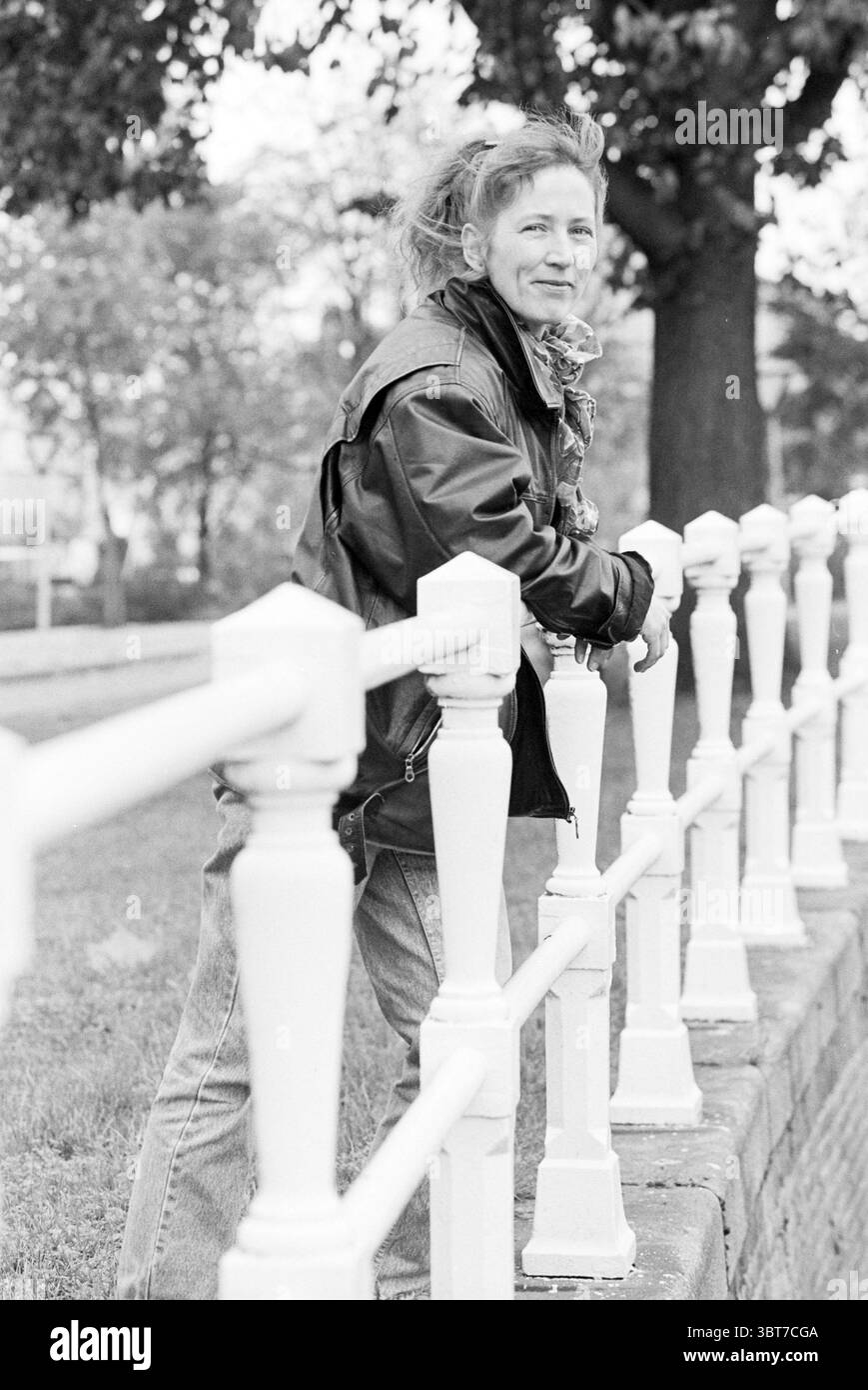 Joke Hofstra; Jugend und Familie, Whizgle News, Dutch Desk, Niederlande, 1950 - 2000 am 01-11-1995. Dies sind die Themen im Bild. Die Szene zeigt eine Person, die neben einem weißen Zaun steht, der ordentlich gesäumt ist und einen Kontrast zum umgebenden Grün bildet. Die Person trägt eine dunkle Lederjacke und eine lässige Jeans aus Denim, die einen lässigen und doch stylischen Look verleiht. Ihr Haar ist leicht zerzaust, was auf eine Bewegung hindeutet, möglicherweise durch eine sanfte Brise. Der Hintergrund ist mit weichen Bäumen gefüllt, deren Blätter verschiedene Grüntöne zeigen, während der Boden ein lebendiger Pa ist Stockfoto