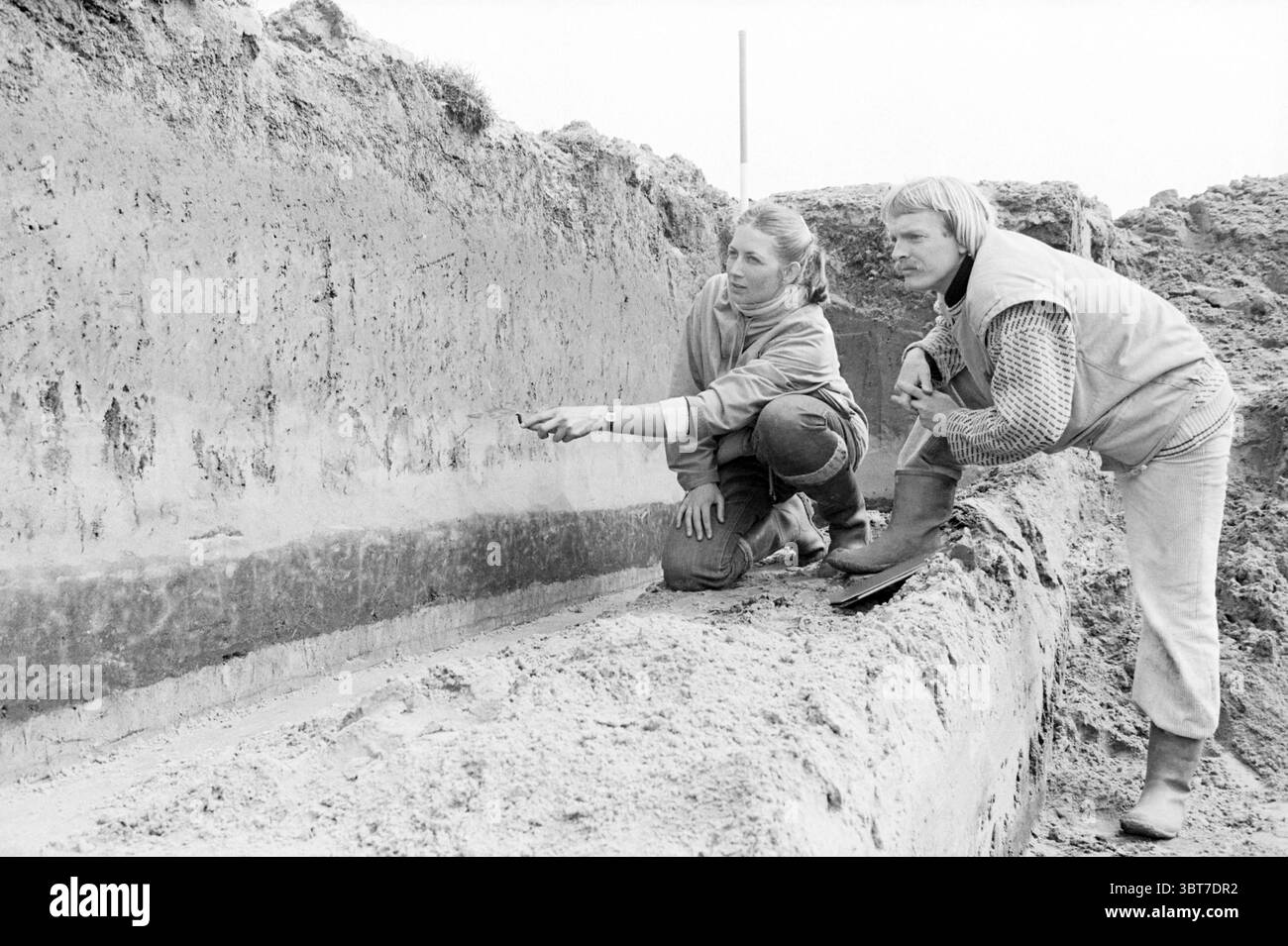 Archäologische Ausgrabungen Velserbroekpolder Ausgrabungen, Whizgle News, Dutch Desk, Niederlande, 1950 - 2000 am 16-04-1985. Dies sind die Elemente im Bild. In einer sonnendurchfluteten Ausgrabungsstätte im Freien werden zwei Personen mit einer detaillierten Analyse eines Grabens beschäftigt, der in die Erde geschnitten wurde. Die Umgebung bietet unebene Erdhügel mit einem mehrschichtigen Erscheinungsbild, die verschiedene Bodenstrukturen und -Farben freigeben, von dunklen bis helleren Tönen. Das erste Individuum wird auf einem Knie gehockt und zeigt auf einen Abschnitt des freiliegenden Bodens. Sie trägt ein langärmliges Hemd und hat ihre Haare zurück gebunden, ich Stockfoto
