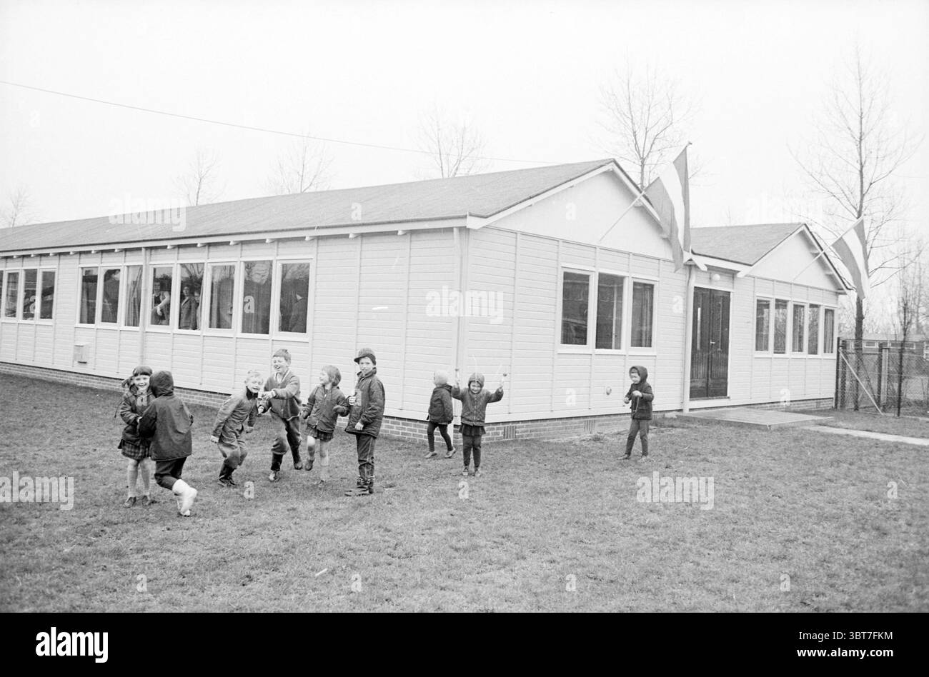 Jugendliche spielen vor zwei Fußballkantinen..., Whizgle News, Dutch Desk, Niederlande, 1950 - 2000. Das Bild enthält diese Themen. Die Szene zeigt eine lebhafte Außenumgebung vor einem bescheidenen, einstöckigen Gebäude, das von einer hellen Holzfassade geprägt ist. Das Gebäude verfügt über zahlreiche Fenster, die einen Einblick in das Innere ermöglichen, und hat ein einfaches Giebeldach mit zwei Flaggen, die auf das Vorhandensein einer Schule oder eines Gemeindezentrums hinweisen. Eine Gruppe von Kindern, die in Wintermänteln und warmer Kleidung gekleidet sind, engagiert sich aktiv im Spiel. Ihre Ausdrücke zeigen Freude und Begeisterung wie sie Stockfoto