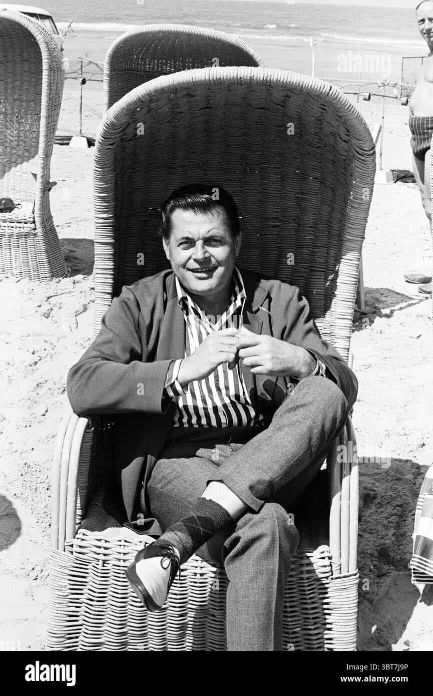 Menschen am Strand Scheveningen, Whizgle News, Dutch Desk, Niederlande, 1950 - 2000 am 09-06-1964. Das Bild enthält diese Themen. Ein Mann sitzt bequem in einem großen, gewebten Liegestuhl, dessen hoher Rücken deutlich hinter ihm steht. Der Stoff seines gestreiften Hemdes steht im Kontrast zu den neutralen Tönen des Stuhls und verleiht der Szene einen stilvollen Touch. Seine entspannte Haltung, mit einem über das andere gekreuzten Bein, deutet auf Leichtigkeit und Zuversicht hin. Im Hintergrund erstreckt sich der Sandstrand zum Meer hin, wo man sanfte Wellen am Ufer beobachten kann. Der Horizont ist klar und zeigt eine sonne an Stockfoto