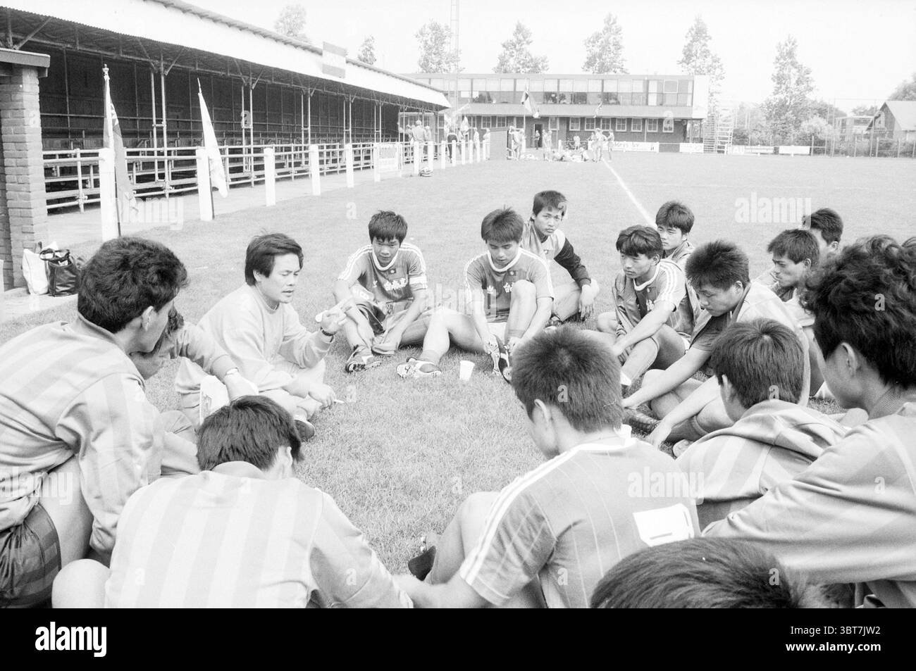 Chinesische Fußballspieler schießen Fußball, Whizgle News, Dutch Desk, Niederlande, 1950 - 2000 am 13-08-1988. Dies sind die Themen im Bild. Auf einem grasbewachsenen Feld, das von einem robusten Gebäude umgeben ist, versammelt sich eine Gruppe junger Athleten in einem engen Kreis am Boden. Die Jungen in passenden Sportuniformen zeigen eine Farbmischung, überwiegend in hellen Blau- und Rottönen, wobei einige Trikots ihre Teamzugehörigkeit hervorheben. Das Schauspiel ist lebhaft, aber konzentriert und spiegelt die Kameradschaft und Entschlossenheit der Spieler wider. Sie sitzen eng zusammen und lehnen sich innig wie die Stockfoto