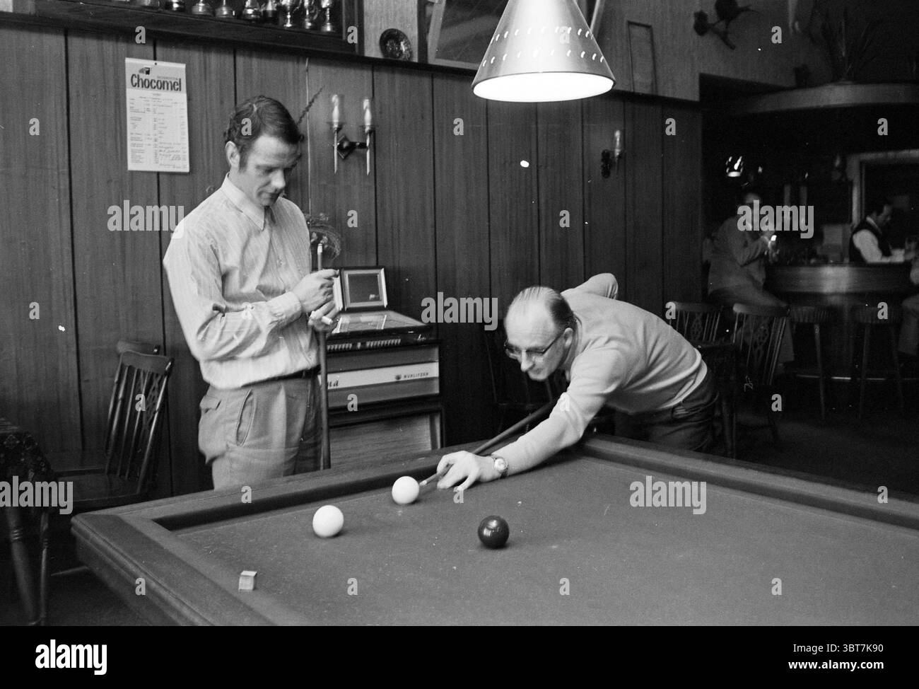 Billard im Café de Zon spielen. Hochofen-Schachturnier, Whizgle News, Dutch Desk, Niederlande, 1950 - 2000 am 14-01-1971. Diese Themen werden in der Abbildung angezeigt. Die Szene befindet sich in einer gemütlichen Bar im Retro-Stil, die sich durch die warme, dunkle Holztäfelung und die dezente Beleuchtung auszeichnet, die eine intime Atmosphäre schafft. Zwei Personen, die beide in legeren Mid-Century-Kleidern gekleidet sind, spielen eine Partie Billard. Die erste Person, die aufrecht steht, wirkt kontemplativ, während sie einen Stichstock hält. Sein nachdenklicher Ausdruck wird durch das sanfte Leuchten einer Deckenlampe hervorgehoben. Er trägt ein Light-C Stockfoto