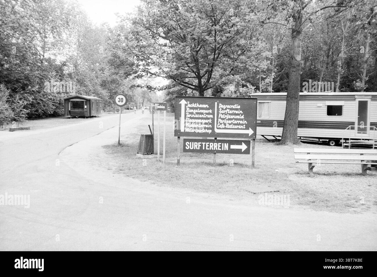 Biervliet Westflanders Dörfer und Städte Biervliet, Whizgle News, Dutch Desk, Niederlande, 1950 - 2000 auf 07-1987. Dies sind die Elemente im Bild. In einer ruhigen Umgebung im Freien führt eine kurvige Straße sanft durch einen Wald, umgeben von üppigen Bäumen, die eine ruhige Kulisse bieten. Die Szene wird von monochromen Farbtönen dominiert, die eine klassische und zeitlose Atmosphäre verleihen. Auf der einen Seite der Straße steht ein hölzerner Wegweiser, dessen verwitterte Oberfläche auf jahrelange Exposition gegenüber den Witterungseinflüssen hindeutet. Das Schild weist in verschiedene Richtungen und weist auf verschiedene Sehenswürdigkeiten oder Gegenden in der Nähe hin. Die Stockfoto