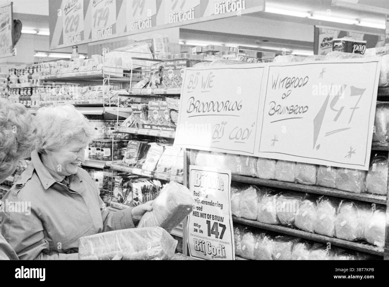 Bread war CODI Market Bread Shops and Kiosks, Whizgle News, Dutch Desk, Niederlande, 1950 - 2000 am 09-11-1982. Diese Themen sind in der Abbildung dargestellt. Die Szene entfaltet sich in einem Lebensmittelbereich, der durch helle, künstliche Beleuchtung gekennzeichnet ist, die eine gut beleuchtete Atmosphäre schafft. Eine Frau, die ein warmes Lächeln zeigt, steht vor einer Ausstellung verpackter Waren. Ihr Haar ist in weichen Locken gestylt und sie trägt eine helle Jacke, die ihr fröhliches Auftreten ergänzt. Die Regale um sie herum sind mit ordentlich angeordneten Produkten ausgestattet, darunter verschiedene Brotsorten und andere typische Artikel Stockfoto