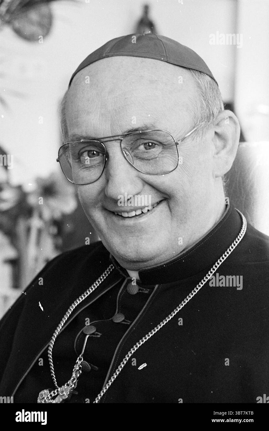 Bischof Bomers Roman Catholic Church, Whizgle News, Dutch Desk, Niederlande, 1950 - 2000 am 23-02-1989. Dies sind die Themen im Bild. Die Szene zeigt einen älteren Mann mit einem warmen und einladenden Ausdruck, der von einem breiten Lächeln geprägt ist, das sein nahes Verhalten unterstreicht. Er hat ein kurzes, helles Haar und trägt eine große runde Brille, die seine freundlichen Augen betont. Seine Kleidung besteht aus dunkler, formeller kirchlicher Kleidung, typisch für ein hochrangiges Mitglied des Klerus. Auf seinem Kopf sitzt eine markante rote Kappe, die seine Position angibt, während eine silberne Kette elegant um den h hängt Stockfoto