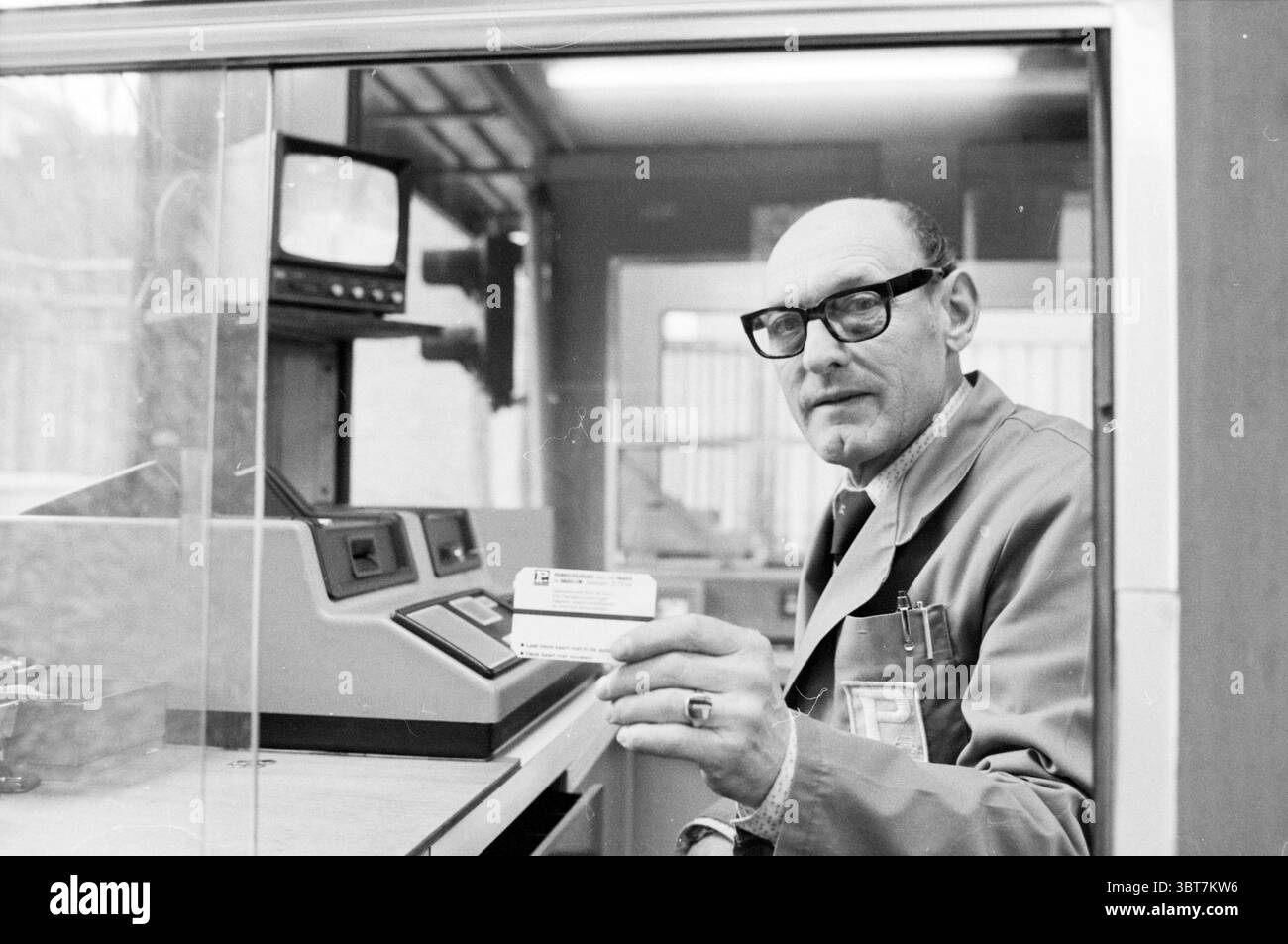 Mann in Raaks Parkhaus mit Parkschein Haarlem Raaks Niederlande, Whizgle News, Dutch Desk, Niederlande, 1950 - 2000 am 01-1982. Das Bild enthält diese Themen. Die Szene zeigt einen älteren Mann, der hinter einer verglasten Kabine sitzt und eine Umgebung andeutet, die ein Tor oder eine Sicherheitsstation sein könnte. Er trägt eine große, dunkle Brille, die seine ausdrucksstarken Augen betont, und einen hellen Mantel, der seinem Auftreten einen professionellen Ton verleiht. Sein sanftes Lächeln strahlt Wärme und Zugänglichkeit aus, während er eine kleine Karte hält, die wahrscheinlich zur Identifizierung oder zum Zugang dient. Der Hintergrund besteht aus Stockfoto