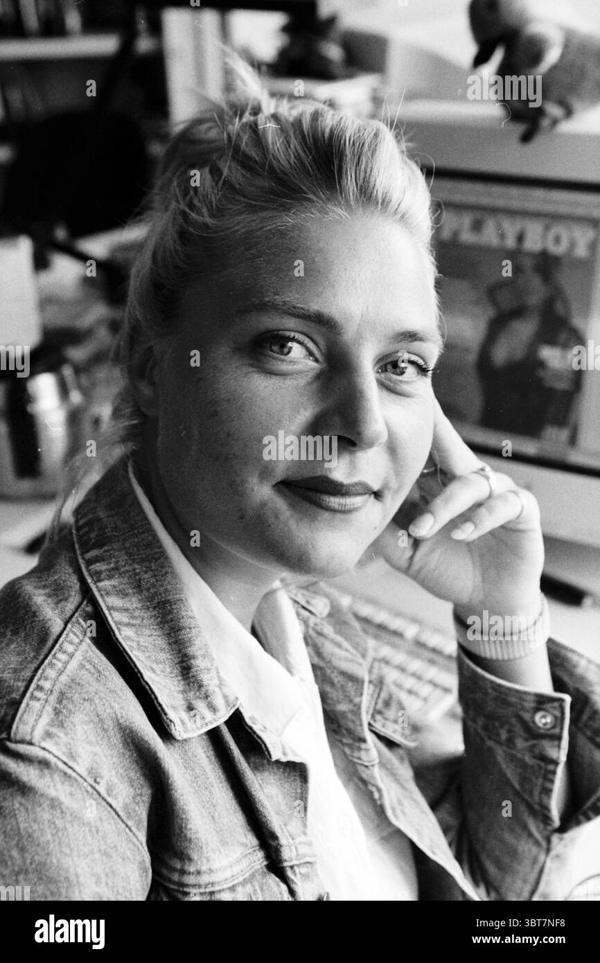 Marian den Toorn Playboy, Whizgle News, Dutch Desk, Niederlande, 1950 - 2000 am 27-08-1997. Dies sind die Elemente im Bild. In einem sanft beleuchteten Innenraum sitzt eine Frau, die ein nachdenkliches, entspanntes Auftreten ausstrahlt. Ihr blondes Haar, leicht zerzaust, umrahmt ihr Gesicht und hebt ihre ausdrucksstarken blauen Augen und ein sanftes Lächeln hervor. Sie trägt eine Denim-Jacke über einem weißen Hemd, was ihrem Aussehen einen lässigen, aber stilvollen Touch verleiht. Im Hintergrund deuten verschiedene Objekte auf einen kreativen Raum hin. Es gibt Anklänge an farbenfrohe Dekor, mit einem Magazin mit einem markanten Cover, das auf ein verspieltes an hindeutet Stockfoto