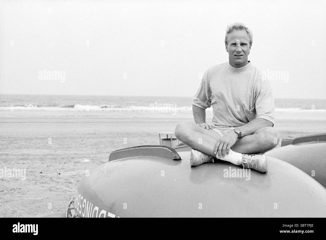 Larry WK Jitskie Free Style, Whizgle News, Dutch Desk, Niederlande, 1950 - 2000 am 09-08-1990. Dies sind die Elemente im Bild. In einer Küstenszene sitzt ein Mann bequem auf der abgerundeten Oberfläche eines leuchtend roten Rettungsbootes, das an einem Sandstrand positioniert ist. Er trägt ein helles kurzärmeliges T-Shirt und sportliche Shorts, kombiniert mit weißen Sneakern, die ein entspanntes und lässiges Auftreten ausstrahlen. Seine Haltung ist im Kreuz, was auf Leichtigkeit und Selbstvertrauen hindeutet, mit einem leichten Lächeln auf seinen Lippen. Die Kulisse bietet einen ausgedehnten Strandabschnitt mit sanften Wellen, die auf dem liegen Stockfoto