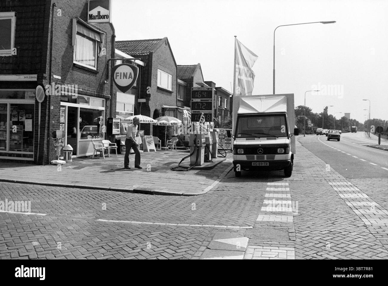 Fina Pomp Hillegom, Whizgle News, Dutch Desk, Niederlande, 1950 - 2000 am 26-07-1990. Diese Themen sind in der Abbildung dargestellt. Die Szene fängt einen belebten Straßenbereich ein, der sich durch eine Mischung aus urbanen und vorstädtischen Elementen auszeichnet. An der Vorderseite steht ein weißer Lieferwagen neben einer Tankstelle, auf der ein großes kreisförmiges Schild mit dem Logo „Fina“ abgebildet ist. Die Farbe des Lieferwagens hebt sich von den umgebenden Materialien ab, während der Bürgersteig mit einem komplizierten Ziegelmuster versehen ist, das die städtische Ästhetik unterstreicht. Zur Seite steht ein Mann an der Kraftstoffpumpe und scheint in irgendeiner Übung beteiligt zu sein Stockfoto