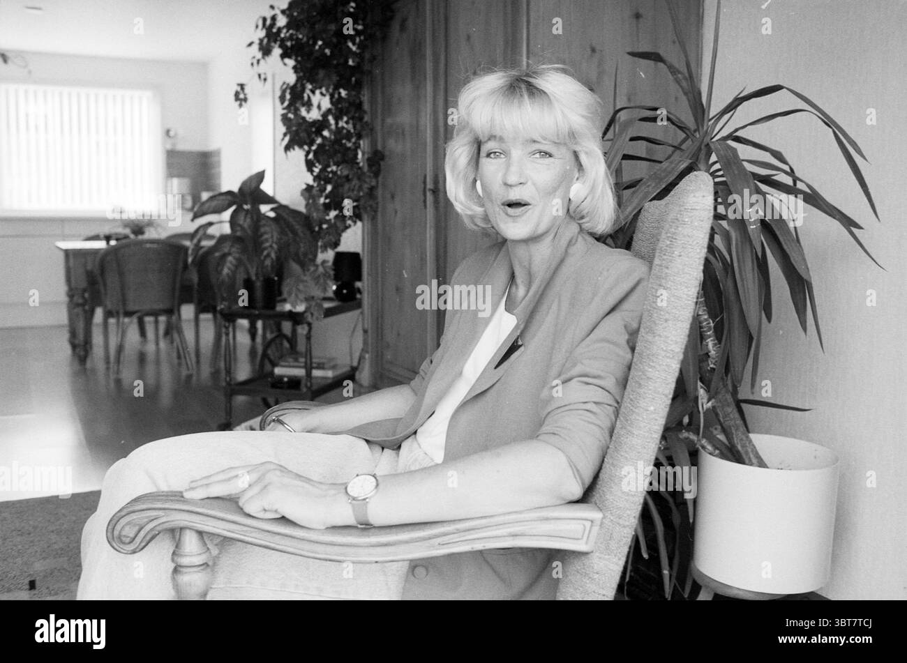 Porträt der Dame im Stuhl, Whizgle News, Dutch Desk, Niederlande, 1950 - 2000 im Jahr 1991. Das Bild enthält diese Themen. In einem warm beleuchteten Innenraum sitzt eine Frau bequem in einem großen, gepolsterten Sessel. Sie hat hellblonde Haare, die in sanften Wellen gestylt sind, und ist in einem hellen Ensemble gekleidet, das eine lässige und dennoch stilvolle Atmosphäre verleiht. Ihr Ausdruck ist lebendig und einnehmend und deutet auf einen Moment des Lachens oder Gesprächs hin. Die Umgebung umgibt sie mit einer Mischung aus natürlichen und künstlichen Elementen. Hinter ihr lassen die großen Fenster das Tageslicht durchleuchten und erleuchten den Raum Stockfoto