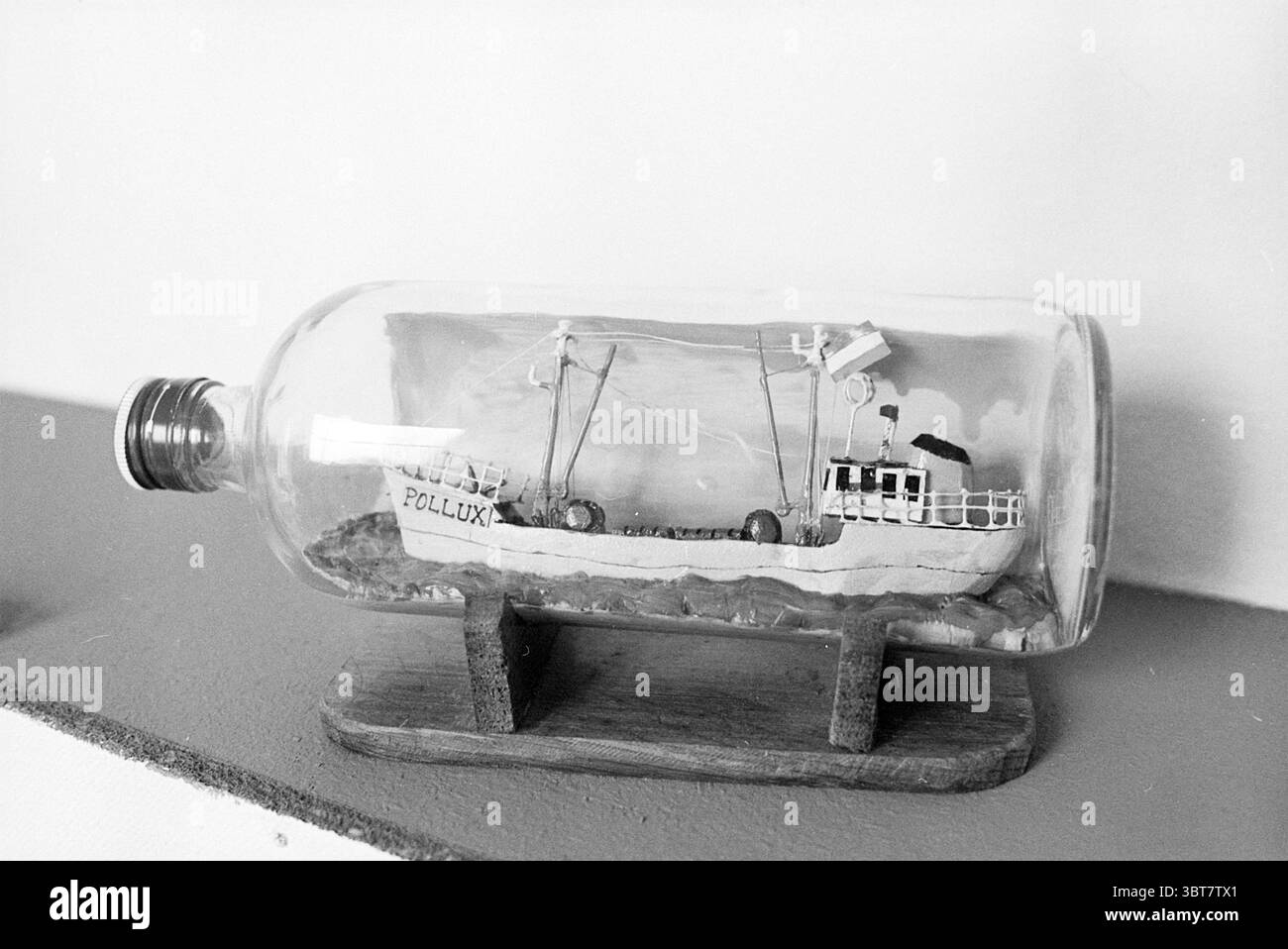 Miniaturboot in Flasche, Whizgle News, Dutch Desk, Niederlande, 1950 - 2000 im Jahr 1977. Diese Themen werden in der Abbildung angezeigt. Die Szene zeigt eine durchscheinende Glasflasche, die auf einem einfachen Holzsockel sichtbar ist. In der Flasche befindet sich ein sorgfältig gefertigtes Miniaturboot namens „Pollux“. Das Boot ist in weiß- und Schwarztönen lackiert und zeigt komplizierte Details wie einen Schornstein und kleine Fahnen, die in einer fiktiven Brise flattern. Um das Boot herum ist eine handgefertigte Meereslandschaft mit strukturierten Elementen dargestellt, die Wellen darstellen und der Komposition Tiefe verleihen. Die Gesamtfarbpalette ist Mist Stockfoto
