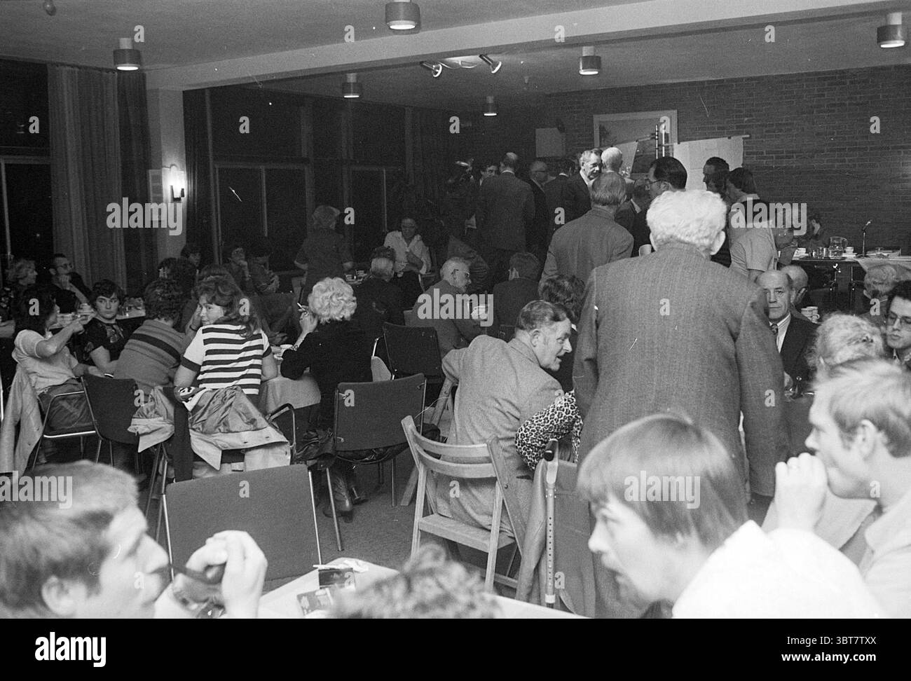 Meeting Old IJmuiden (Gemeinde Breezicht IJmuiden) Meetings, Whizgle News, Dutch Desk, Niederlande, 1950 - 2000 am 14-10-1977. Dies sind die Elemente im Bild. Die Szene zeigt ein lebhaftes Treffen in einem geräumigen, gut beleuchteten Raum mit verschiedenen Sitzmöglichkeiten. Die Atmosphäre ist voll von Gesprächen und Interaktionen unter den Teilnehmern. Die Menschen führen animierte Diskussionen, einige sitzen an Tischen, andere bewegen sich durch die Stühle und Gruppen. Die Teilnehmer scheinen ein unterschiedliches Alter zu repräsentieren, von jungen Erwachsenen bis hin zu reiferen Erwachsenen Stockfoto