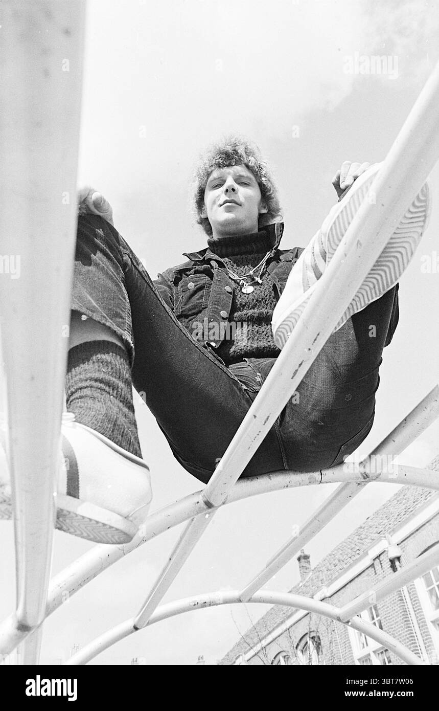 Porträt von Floor de Vos Porträts, Whizgle News, Dutch Desk, Niederlande, 1950 - 2000 am 28-04-1970. Diese Themen werden in der Abbildung angezeigt. Die Szene zeigt eine Person, die auf einer Metallkonstruktion sitzt, vielleicht ein Spielplatz oder eine städtische Installation. Die Person ist in einem Winkel positioniert, der ihr entspanntes Verhalten betont, wenn sie nach oben blickt und ein beiläufiges Selbstvertrauen ausstrahlt. Ihre Kleidung besteht aus einer dunklen Denim-Jacke und einem Strickpullover, was einen bequemen und doch stylischen Look vermittelt. Auch die Jeans sind dunkel und ergänzen ihr Outfit. Zu den Schuhen gehören wahrscheinlich weiße Sneakers Stockfoto