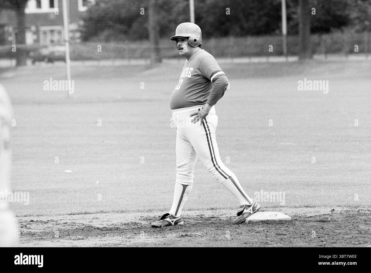 Softball Männer: Bloemendaal-Almere Bloemendaal, Whizgle News, Dutch Desk, Niederlande, 1950 - 2000 am 22-06-1991. Diese Themen sind in der Abbildung dargestellt. Die Szene zeigt einen Baseballdiamanten unter einem bewölkten Himmel, was auf einen bewölkten Tag hindeutet. Das Gras, das das Feld umgibt, ist ein leuchtendes Grün, sorgfältig gepflegt und dennoch weich unter den Füßen. Im Vordergrund steht ein Spieler selbstbewusst auf einer Basis. Er trägt ein tailliertes, kurzärmeliges Trikot, dazu eine helle Hose mit einem einzelnen Streifen an der Seite. Das Trikot ist in einem tiefen Farbton gehalten, der sich stark von seiner Kleidung abhebt. Seine Rundung Stockfoto