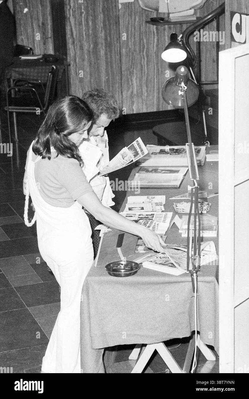 Open Day Konzert Hall Publikum mit Broschüren Haarlem the Netherlands, Whizgle News, Dutch Desk, Niederlande, 1950 - 2000 am 01-09-1979. Diese Themen sind in der Abbildung dargestellt. In einem belebten Innenbereich stehen zwei Personen eng zusammen und untersuchen verschiedene gedruckte Materialien auf einem Tisch. Die Frau, gekleidet in einem taillierten weißen Outfit, lehnt sich leicht nach vorne und zeigt auf etwas Interessantes, während sie ein Hochglanzmagazin in der anderen Hand hält. Ihr dunkles Haar fällt in sanfte Wellen, bedeckt teilweise ihre Schultern, und sie strahlt einen Hauch von Neugier und Begeisterung aus. Der Mann Be Stockfoto