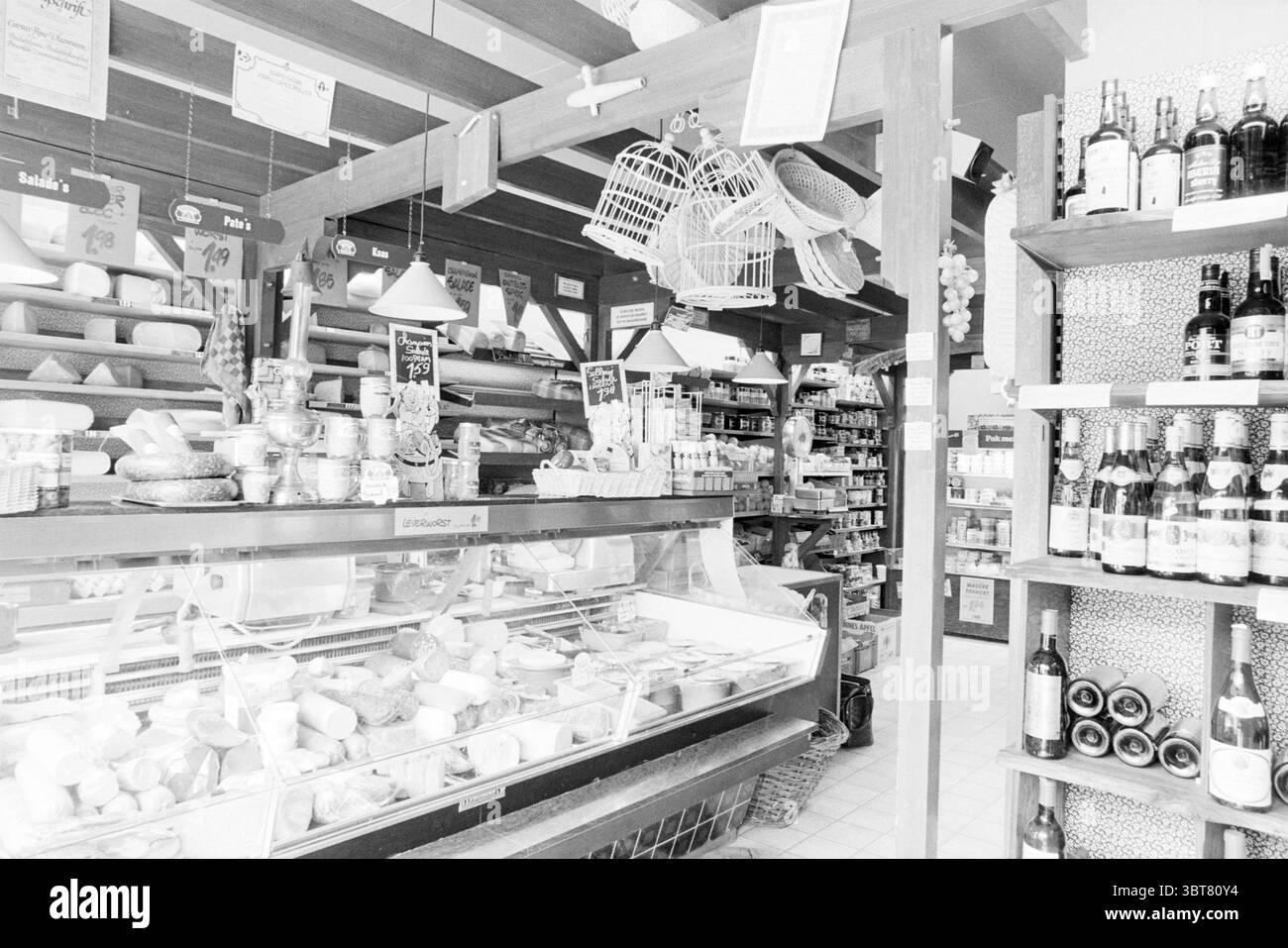 Kaufen Sie den Lebensmittelhändler Aad van der Peet mit Brot, Milchprodukten und Fleisch an der Ecke Borneostraat und Celebesstraat mit dem Eingang auf Borneostraat Heemstede. Geschäfte und Kioske. Heemstede Borneostraat the Netherlands, Whizgle News, Dutch Desk, Niederlande, 1950 - 2000 auf 30-06-1980. Diese Themen werden in der Abbildung angezeigt. Die Szene präsentiert einen hübschen Supermarkt mit rustikalem Charme. Regale mit verschiedenen Waren säumen die Wände und zeigen eine Auswahl an Gläsern, Flaschen und verpackten Artikeln, die sowohl lokale als auch handwerkliche Produkte andeuten. Die Farbpalette ist überwiegend monochrom und unterstreicht das nostalgische A Stockfoto