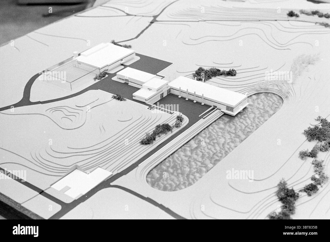 Modell des Gebäudes., Whizgle News, Dutch Desk, Niederlande, 1950 - 2000. Diese Themen sind in der Abbildung dargestellt. Die Szene zeigt ein detailliertes Architekturmodell, das eine moderne Struktur in einer sanft hügeligen Landschaft darstellt. Das Modell ist überwiegend monochromatisch und betont die klaren Linien und geometrischen Formen der Gebäude statt Farben oder Texturen. Im Vordergrund hebt sich ein zentrales Gebäude mit breiten, flachen Flächen und einem minimalistischen Design hervor, das eine zeitgenössische Ästhetik suggeriert. Sein flaches Dach erstreckt sich über eine Reihe rechteckiger Abschnitte, wodurch ein Gefühl von Offenheit entsteht Stockfoto