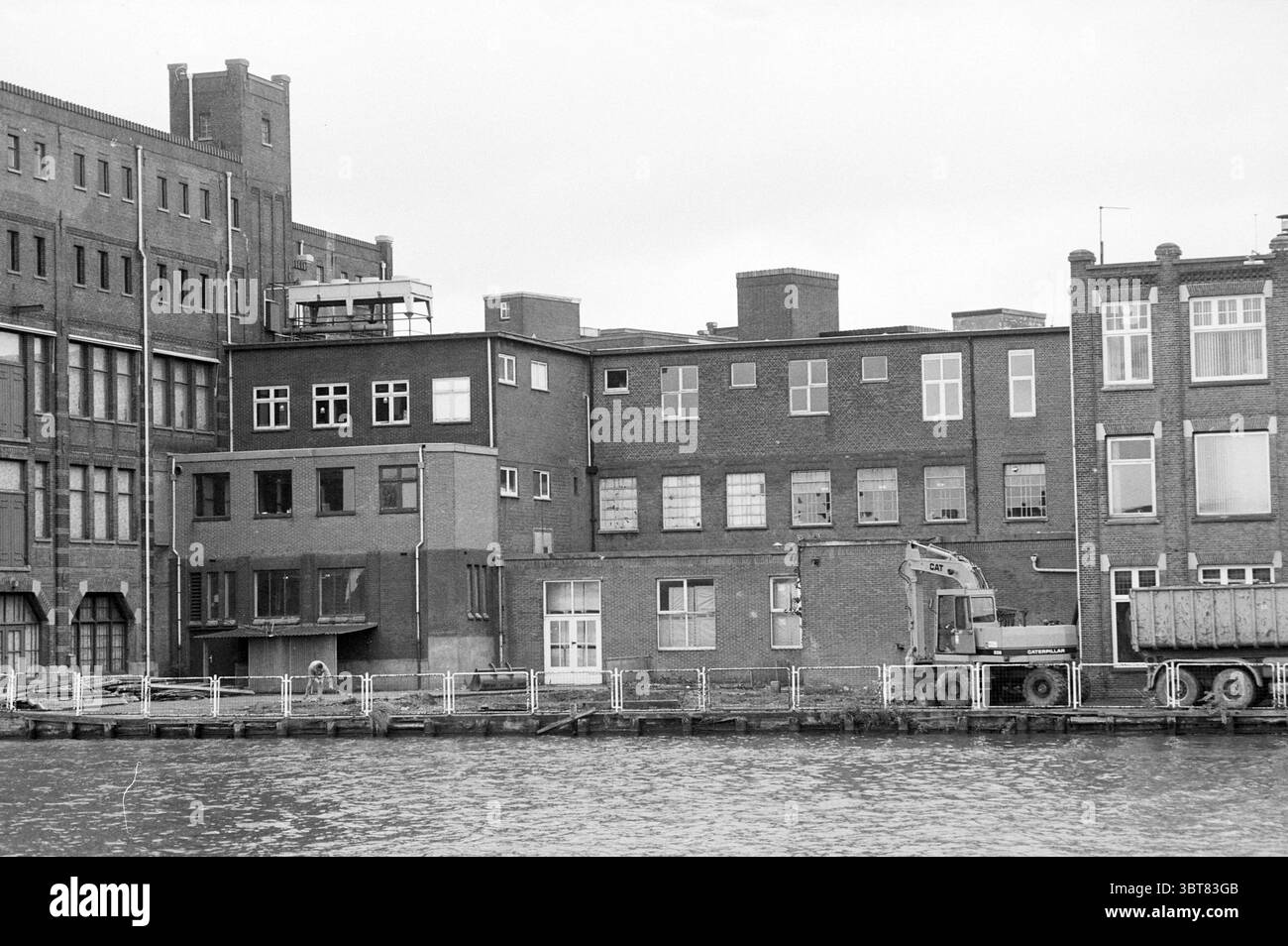 Abriss des Gebäudes in der Nähe von Droste Haarlem, Niederlande, Whizgle News, Dutch Desk, Niederlande, 1950 - 2000 am 09-09-1990. Dies sind die Themen im Bild. Die Szene zeigt eine Uferansicht einer Sammlung von Industriegebäuden. Die Strukturen zeichnen sich durch ihre Ziegelfassaden aus, wobei gedämpfte Grau- und Brauntöne die Palette dominieren. Die Gebäude haben einen imposanten, zweckmäßigen Aspekt, mit unterschiedlich großen Fenstern, die ein scharfes, rechteckiges Muster widerspiegeln. Einige Fenster scheinen zerschlissen oder verschlossen zu sein, was auf Alter und Vernachlässigung hindeutet. Das Wasser davor ist still, mirr Stockfoto