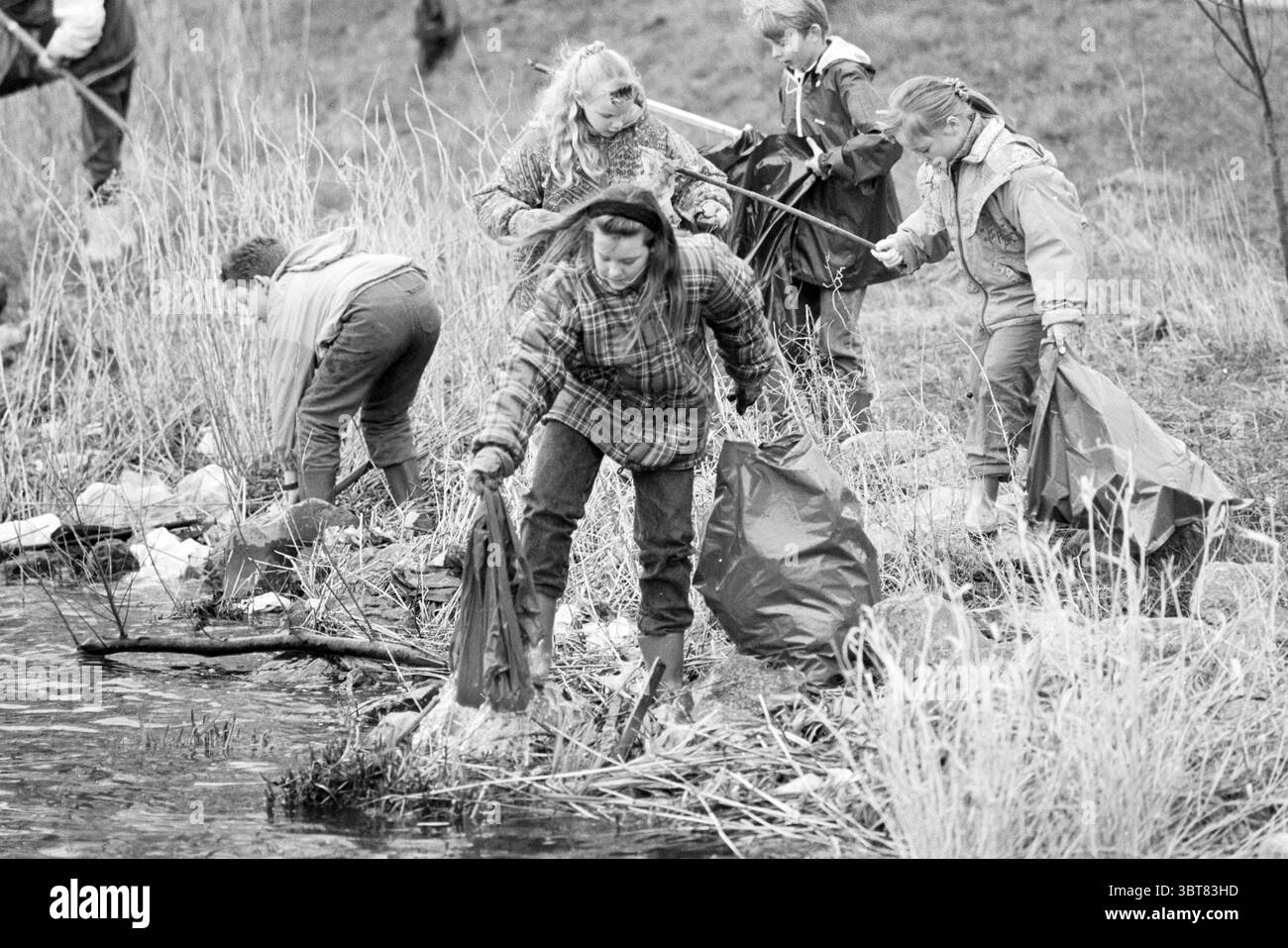 Reinigung von Side Canal C durch einen Studenten, Whizgle News, Dutch Desk, Niederlande, 1950 - 2000 am 20-02-1995. Das Bild enthält diese Themen. Eine Gruppe von Kindern und Teenagern ist an einer Säuberungsaktion entlang des Ufers einer Wasserstraße beteiligt. Der Schwerpunkt liegt auf deren Maßnahmen, während sie Schmutz aus dem Wasser und den umliegenden Bereichen sammeln. Die Szene spielt in einer natürlichen Umgebung mit einer Mischung aus hohen Gräsern, Schilf und kleinen Sträuchern, die den Rand des Wassers umrahmen. Das Wasser wirkt ruhig, aber leicht trüb und spiegelt wahrscheinlich die nahe gelegene Vegetation statt eines klaren Himmels wider. Die Einzelpersonen sind in ungezwungener Outdoor-Kleidung gekleidet Stockfoto