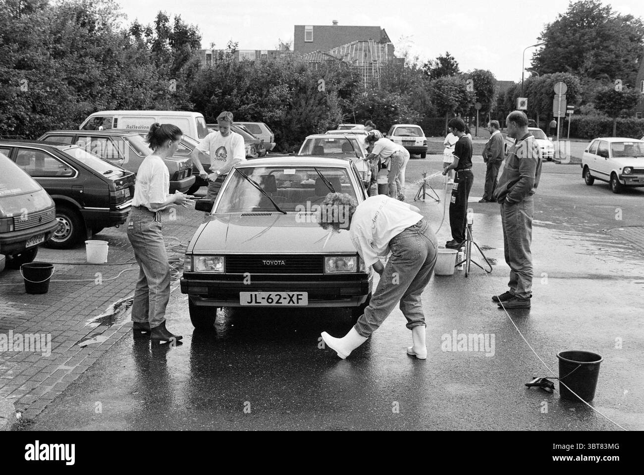 Autowaschanlage Hoofddorp the Netherlands, Whizgle News, Dutch Desk, Niederlande, 1950 - 2000 am 26-05-1990. Diese Themen werden in der Abbildung angezeigt. In einer belebten Outdoor-Szene ist eine Gruppe von Menschen an einer Autowaschanlage beteiligt. Die Umgebung verfügt über einen Parkplatz mit mehreren Fahrzeugen, die unter hellem Himmel stehen, was auf einen sonnigen Tag hindeutet. Im Mittelpunkt steht ein kompakter, zentral positionierter Oldtimer, dessen glänzende Oberfläche von einer Person sorgfältig geschrubbt wird. Diese Person ist lässig gekleidet, trägt helle Kleidung und weiße Gummistiefel, was auf den Eifer hinweist, sich mit ihm auseinanderzusetzen Stockfoto