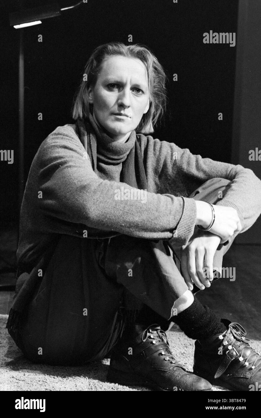 Anneke van Blokland Direktorin, Whizgle News, Dutch Desk, Niederlande, 1950 - 2000 am 12.08.1987. Diese Themen sind in der Abbildung dargestellt. In einem schwach beleuchteten Raum sitzt eine Figur auf dem Boden mit einem komponierten, aber introspektiven Ausdruck. Das Motiv trägt einen lässigen, übergroßen Pullover und eine locker geschnittene Hose und vermittelt ein entspanntes, aber nachdenkliches Verhalten, das durch seine Haltung unterstrichen wird – ein Bein gebeugt und das andere ausgestreckt. Die Umgebung wirkt minimalistisch, mit einer subtilen Textur unter dem Fuß, die eine inszenierte Szene andeutet, vielleicht mit einem hellen Teppich, der sich gegen den dunklen Hintergrund abhebt. Ein Fokus Stockfoto