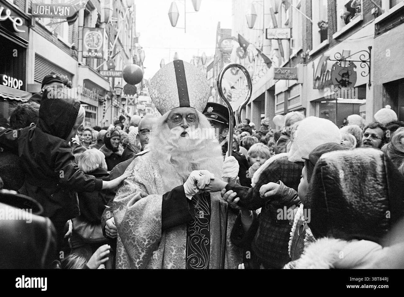 St. Nicholas im Anegang Haarlem St. Nicholas Sinterklaas Haarlem Anegang the Netherlands, Whizgle News, Dutch Desk, Niederlande, 1950 - 2000 am 14-11-1970. Dies sind die Elemente im Bild. In einer lebhaften urbanen Straßenszene herrscht die Atmosphäre vor der Spannung einer festlichen Feier. Eine vielfältige Menschenmenge füllt die enge Straße, mit Erwachsenen und Kindern sichtbar engagiert. Die Szene dominiert eine Figur in einem königlichen, fließenden Gewand, verziert mit satten Rot- und Goldtönen, die Autorität und Tradition symbolisieren. Diese Figur hat einen langen weißen Bart und trägt eine hohe, verzierte Gehrung, Ex Stockfoto