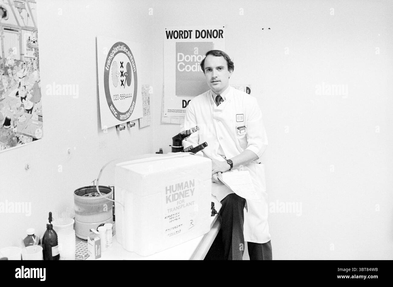 Dr. Strongers zur Nierentransplantation AMC Amsterdam. Medizin Heilmethode Amsterdam Niederlande, Whizgle News, Dutch Desk, Niederlande, 1950 - 2000 am 21-04-1985. Diese Themen sind in der Abbildung dargestellt. Die Szene zeigt einen Mann in einem weißen Labormantel, der selbstbewusst neben einem sauberen, klinischen Arbeitsplatz steht. Seine Kleidung vermittelt Professionalität, ein Hemd und eine Krawatte unter dem Mantel sorgen für einen Hauch von Formalität. Er scheint fokussiert zu sein, blickt direkt auf den Zuschauer und stellt eine unmittelbare Verbindung her. Vor ihm befindet sich ein großer rechteckiger Kühler, der mit dem gekennzeichnet ist Stockfoto
