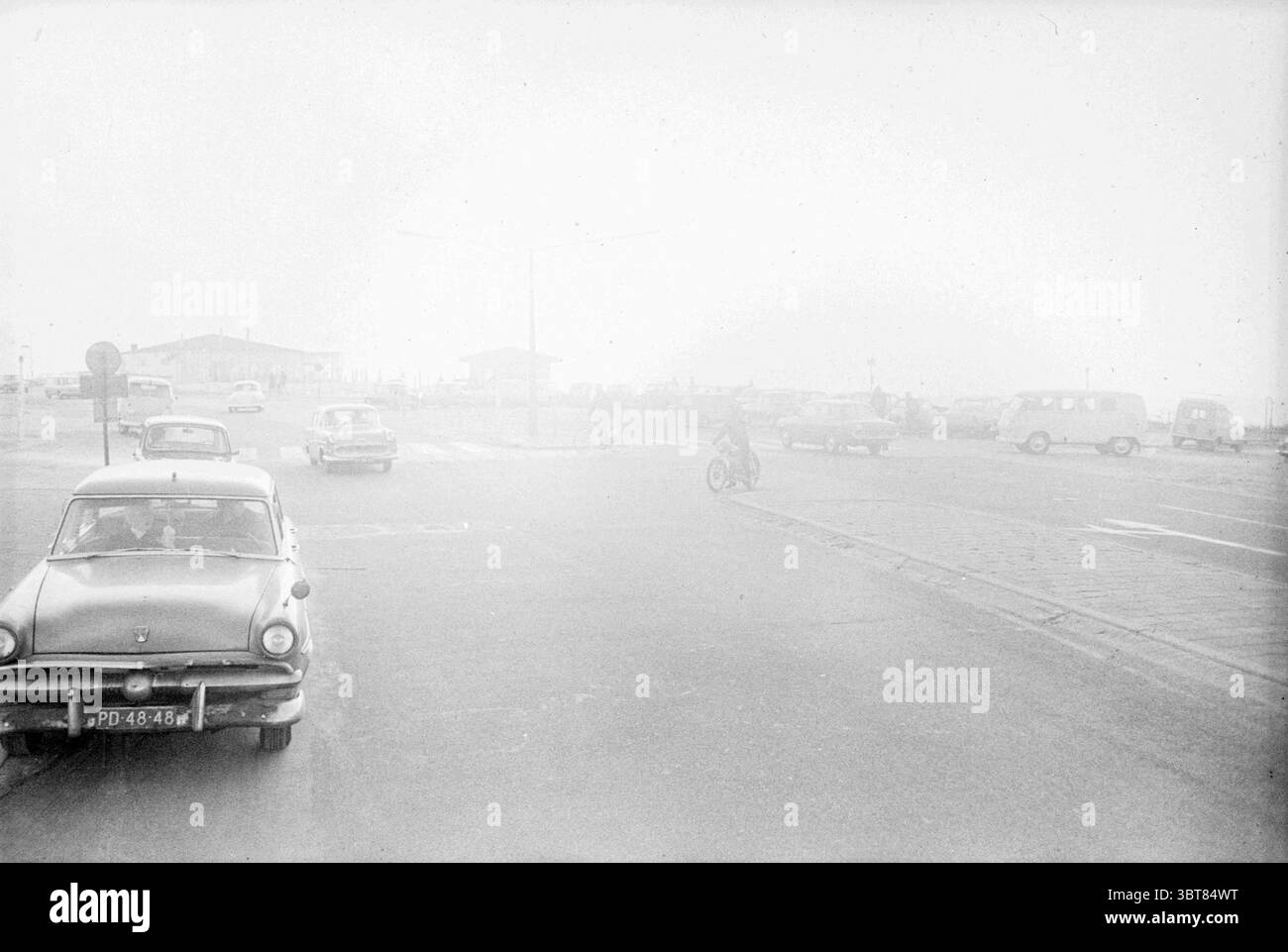 Verkehr auf dem Boulevard, Whizgle News, Dutch Desk, Niederlande, 1950 - 2000 am 14-02-1965. Diese Themen werden in der Abbildung angezeigt. Die Szene ist in Nebel gehüllt, was ihr eine gedämpfte und atmosphärische Qualität verleiht. Die Sichtbarkeit ist eingeschränkt, sodass ein weicher, grauer Farbton über die gesamte Einstellung erzeugt wird. Im Vordergrund befinden sich mehrere Oldtimer, deren klassische Formen durch den Dunst kaum zu erkennen sind. Ein Auto, von der Seite gesehen, hat abgerundete Kanten und eine glänzende Oberfläche, was auf seine Ära hinweist; es scheint an einer Kreuzung geparkt zu sein. Der Fahrer, der durch die Windschutzscheibe sichtbar ist, hat einen kontemplativen Ausdruck. Stockfoto
