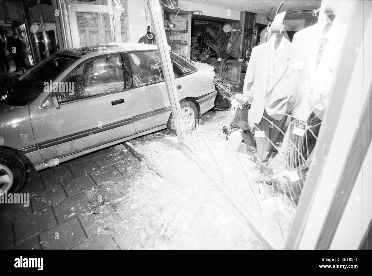 Roka Fashion Robbery Haarlem Julianapark The Netherlands, Whizgle News, Dutch Desk, Niederlande, 1950 - 2000 am 14-11-1991. Dies sind die Elemente im Bild. Die Szene zeigt einen starken Kontrast zwischen Chaos und Ruhe, wobei dunkle Schattierungen die Komposition dominieren. Ein leicht zerknittertes und unpassendes Fahrzeug ist mit einer Ladenfront kollidiert. Das Fahrzeug steht schräg, seine Karosserie weist Dellen auf, die auf einen erheblichen Aufprall hindeuten. Die Schaufensterfront, in der formelle Kleidung wie Anzüge und Krawatten ausgestellt ist, wird teilweise durch zersplittertes Glas verdeckt. Fragmente der Glasstreuung ac Stockfoto