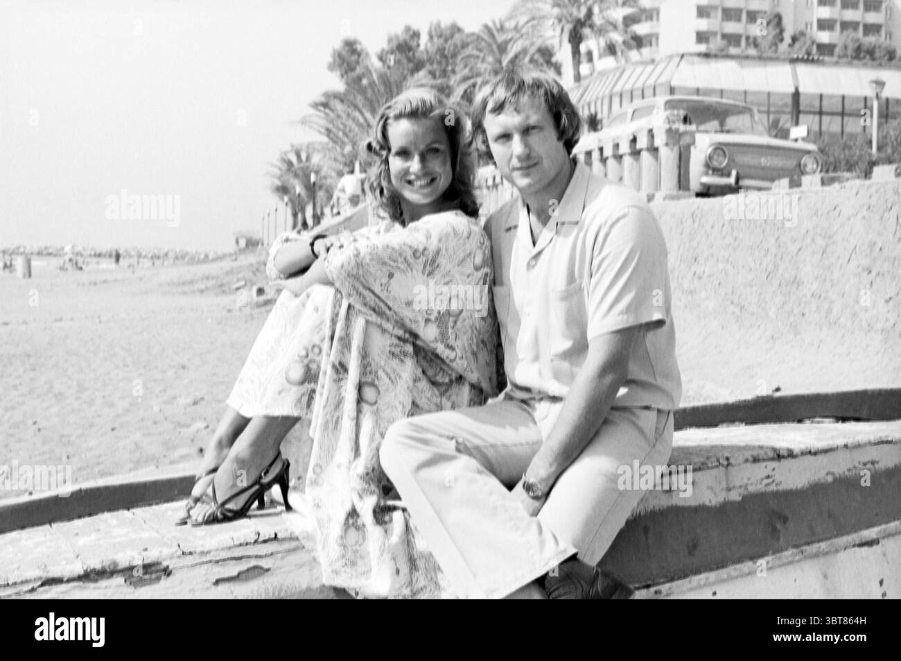 Bericht Torremolinos Spain, Whizgle News, Dutch Desk, Niederlande, 1950 - 2000 am 10-08-1978. Diese Themen sind in der Abbildung dargestellt. In einer Küstenlage posiert ein Paar glücklich an einem Sandstrand. Die Frau, gekleidet in einem fließenden, gemusterten Kleid, hat ein breites Lächeln, das Freude und Wärme verkörpert. Ihr Haar kaskadiert in sanften Wellen und unterstreicht ihr fröhliches Verhalten. Neben ihr sitzt ein Mann in hellem Hemd und Hose bequem. Er neigt sich leicht zu ihr und wirkt entspannt und engagiert. Die Kulisse bietet einen weiten Strand mit einer sanften Welle, die am Ufer plätschert. Handfläche Stockfoto
