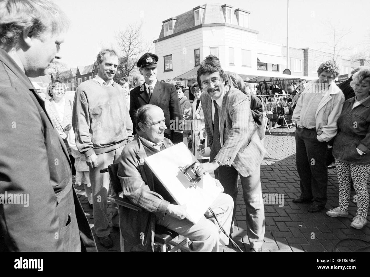 Inbetriebnahme des Strommarktes Florisstraat Inbetriebnahme Haarlem Floresstraat, Whizgle News, Dutch Desk, Niederlande, 1950 - 2000 am 30-03-1989. Die Abbildung zeigt diese Themen. In einer lebhaften Outdoor-Szene versammelt sich eine Gruppe von Menschen, die eine Atmosphäre der Begeisterung und Neugier erzeugen. Die Umgebung scheint ein geschäftiger Markt oder ein öffentlicher Platz zu sein, der von kopfsteingepflasterten Wegen und einer Kulisse malerischer Gebäude mit heller Fassade und schrägem Dach geprägt ist. Im Vordergrund zieht eine sitzende Person Aufmerksamkeit auf sich. Er sitzt auf einem Stuhl des Regisseurs, mit einem warmen Lächeln Stockfoto