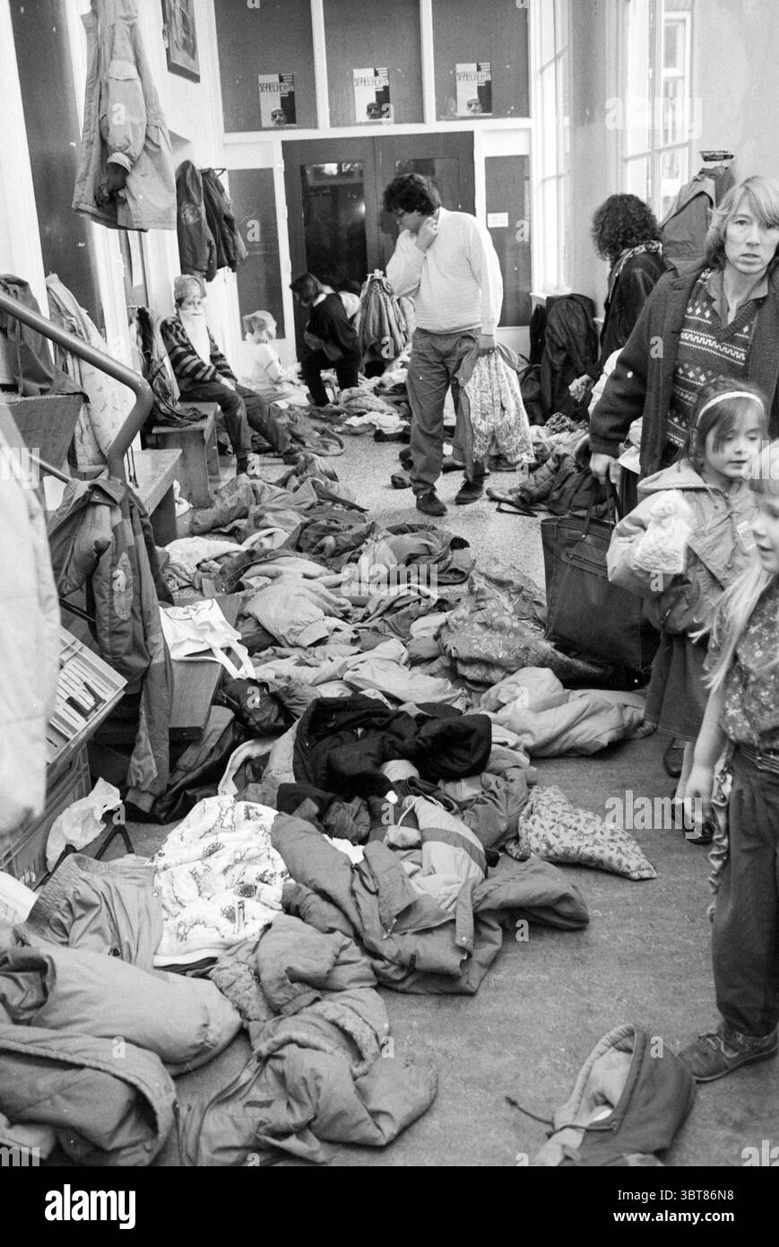 Garderobenständer am GNOME Day, Whizgle News, Dutch Desk, Niederlande, 1950 - 2000 am 01.02.1991. Das Bild enthält diese Themen. Die Szene fängt einen belebten Innenraum ein, wahrscheinlich ein Gemeindezentrum oder ein informeller Treffpunkt, voller Menschen und verstreuter Kleidung. Der Boden wird größtenteils durch eine Reihe von Jacken, Mänteln und verschiedenen Winterkleidung verdeckt, was einen chaotischen Farbteppich und Texturen schafft. Die Kleidungsstücke, hauptsächlich in gedämpften Tönen – Grün, Braun und Grau – wirken abgenutzt, aber funktionell, was darauf hindeutet, dass sie gut verwendet wurden. Im Hintergrund lassen große Glastüren natürliche L Stockfoto
